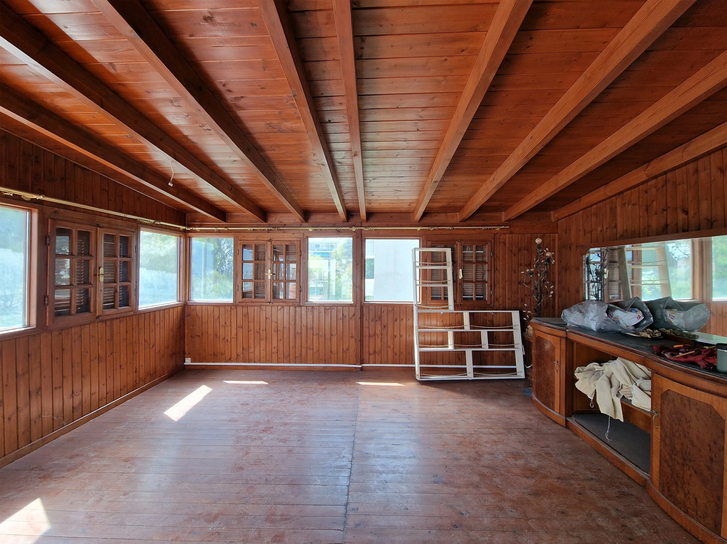 Room with wooden walls and ceiling, large windows, some furniture and items on the counter, and a white storage rack.