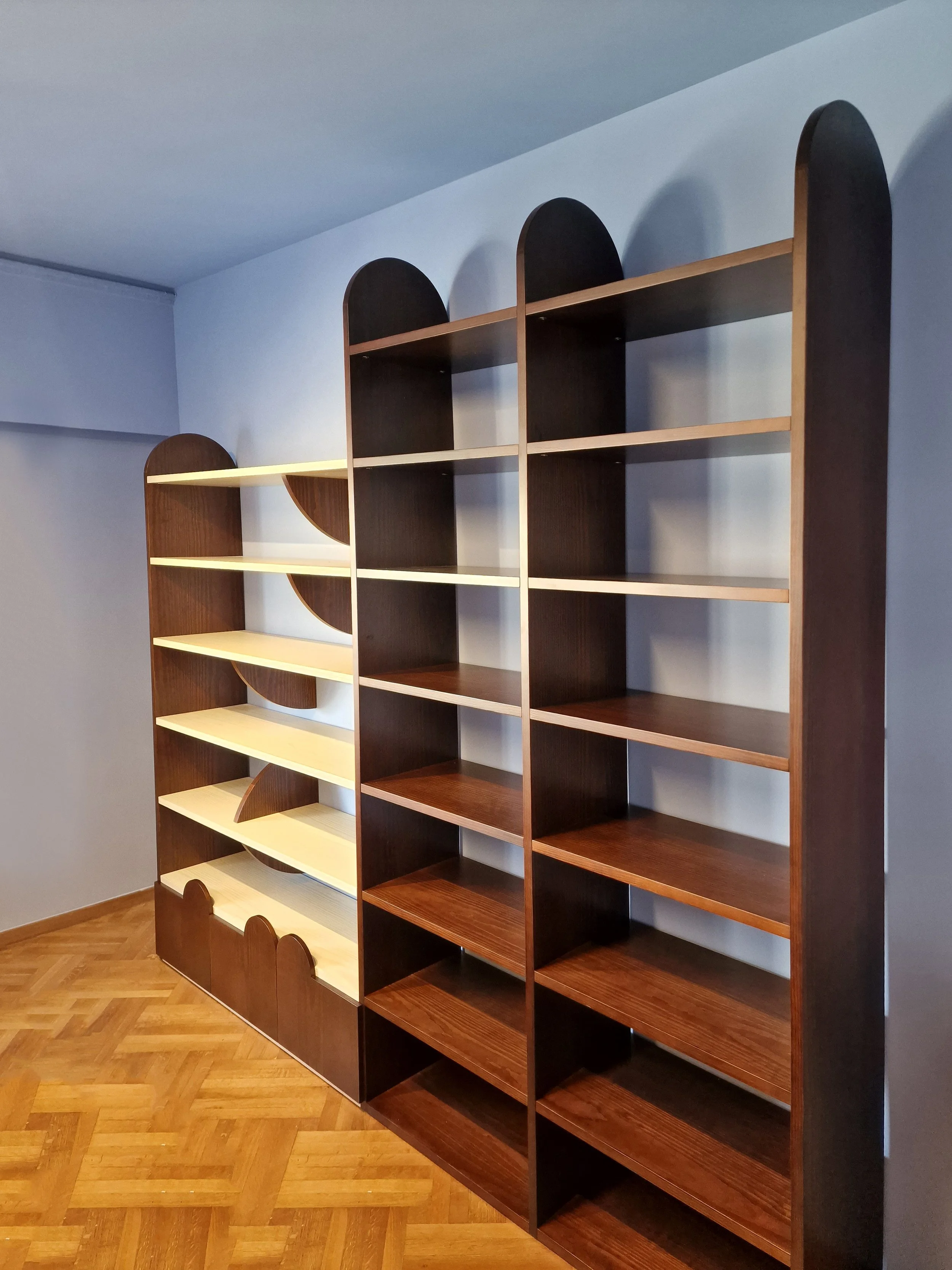 Two wooden bookshelves with arched tops, one dark brown and one light cream, placed in a room with wood parquet flooring and a plain light-colored wall.