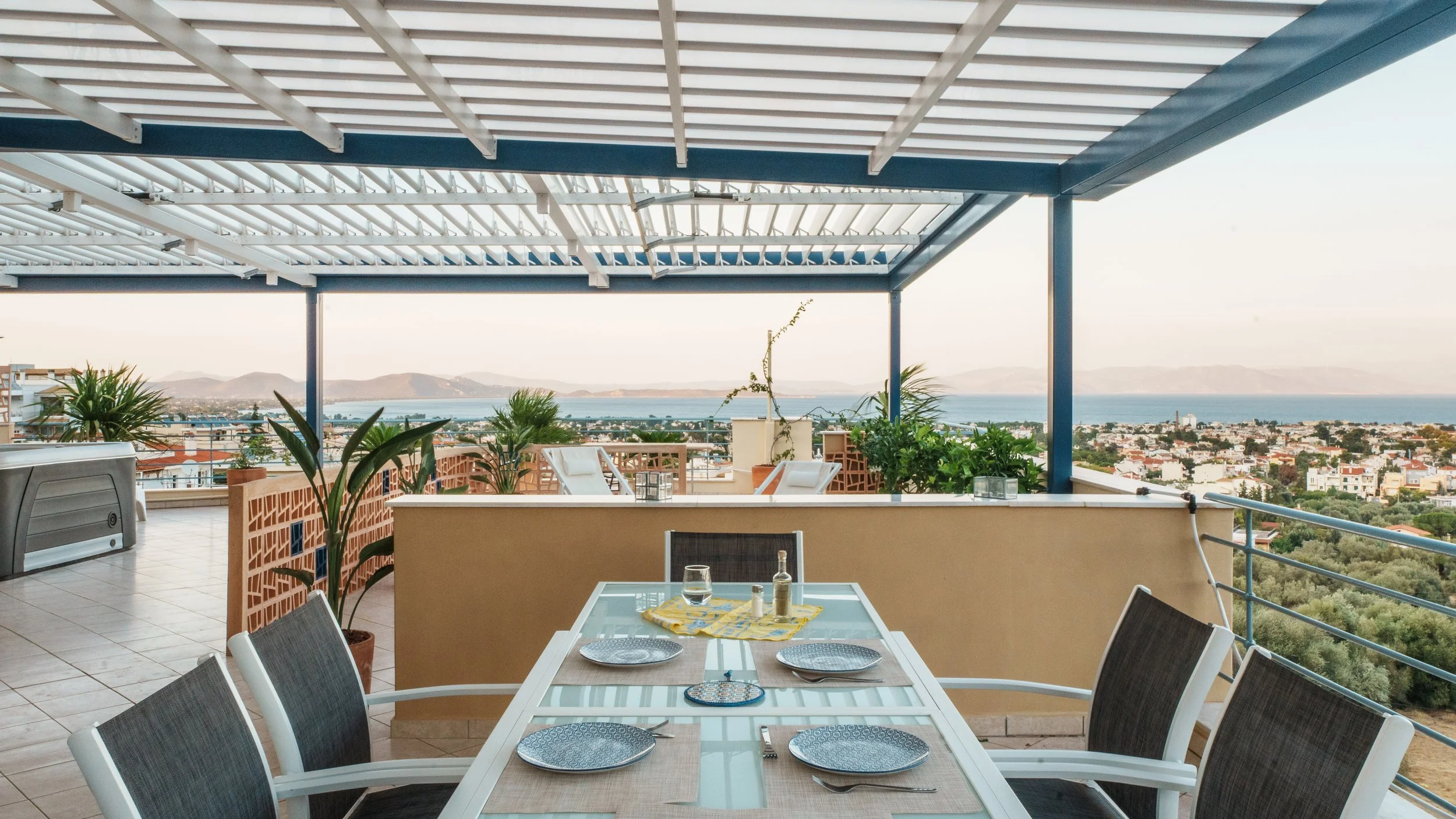 A rooftop terrace with a dining table set with plates, glasses, and utensils, surrounded by chairs, overlooking a cityscape with mountains and water in the distance.