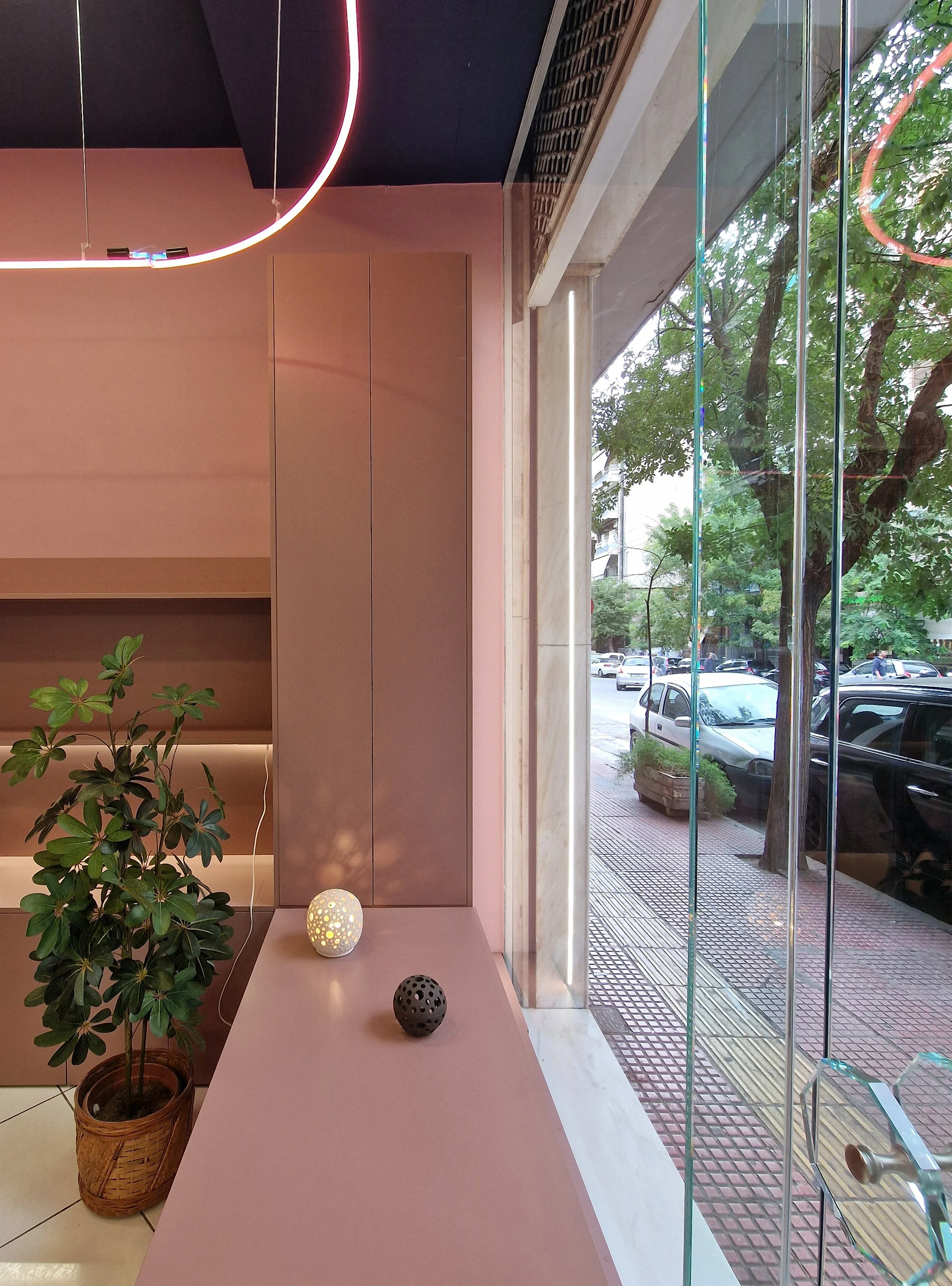 Inside a modern building, a pink counter with a potted plant, decorative spherical lights, and a view of a street with parked cars and trees through large glass windows.