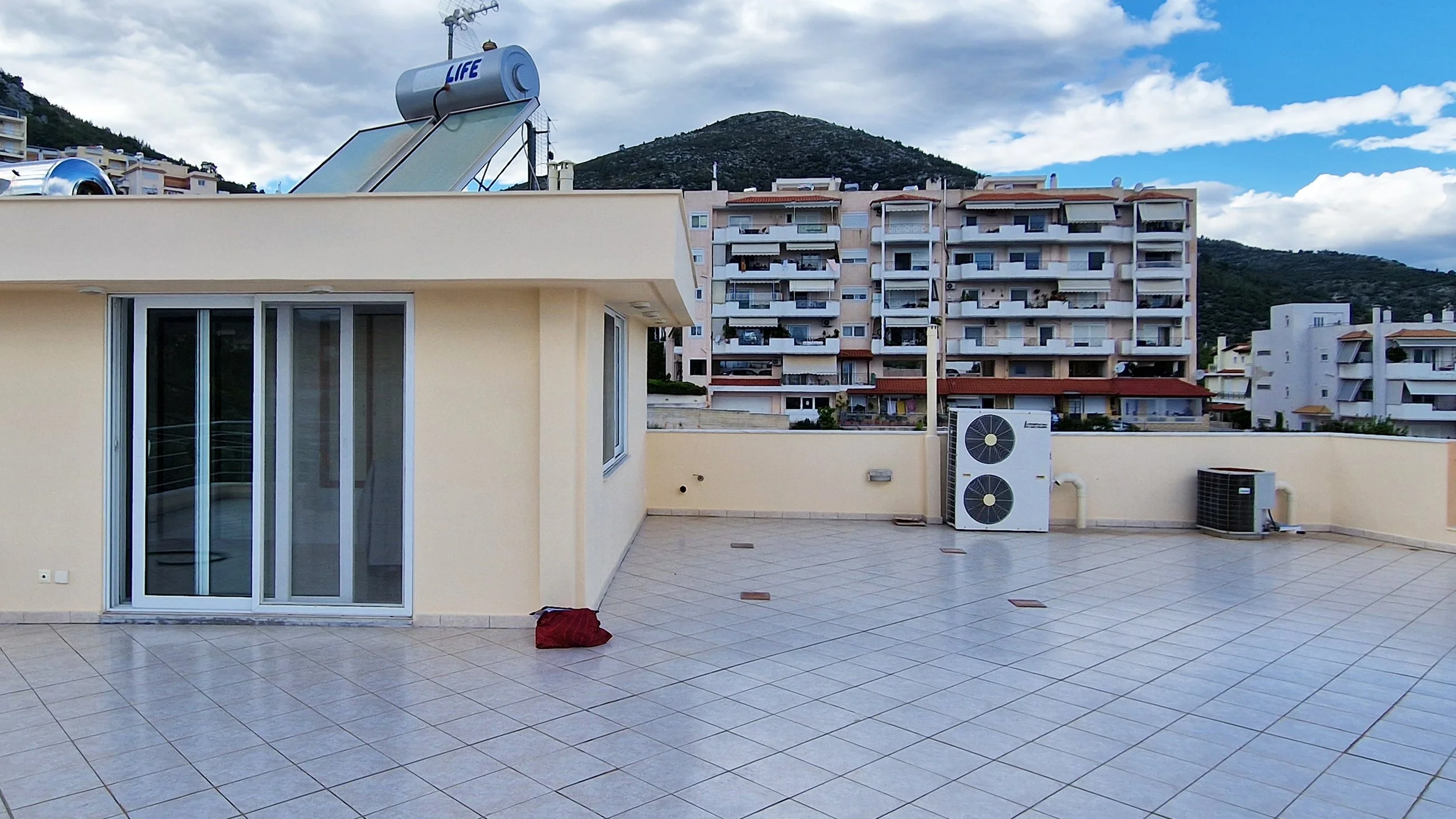 Rooftop terrace with tiled floor, sliding glass door, air conditioning units, and a red cloth or bag on the ground. In the background, there are multiple apartment buildings and a mountain under a partly cloudy sky.