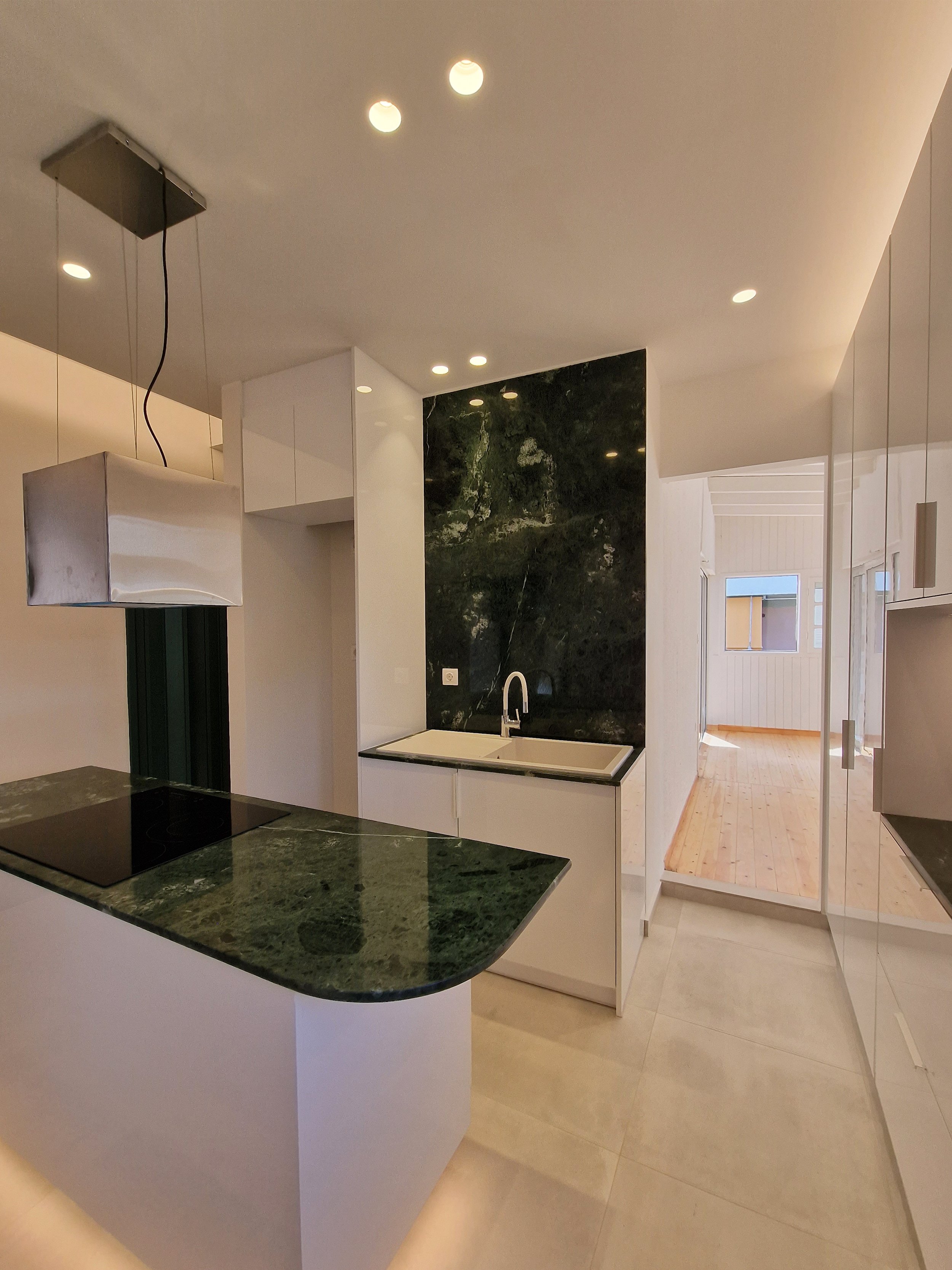 Modern kitchen with white cabinets, black marble backsplash, kitchen sink, green marble island counter, and a view into a sunlit room with wooden floor and windows.