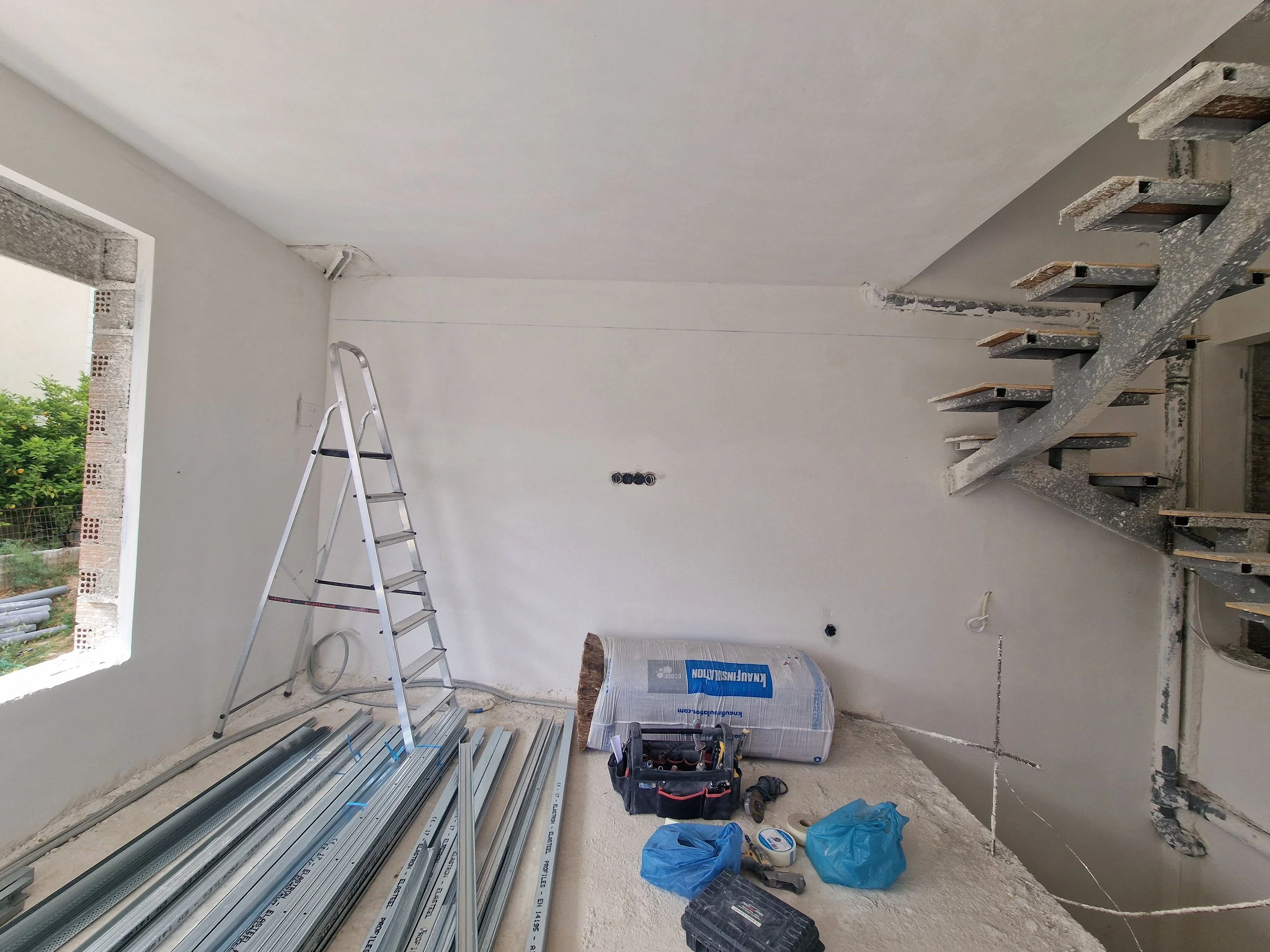 Interior of a construction site with metal framing, a stepladder, building tools, and materials scattered on the floor.