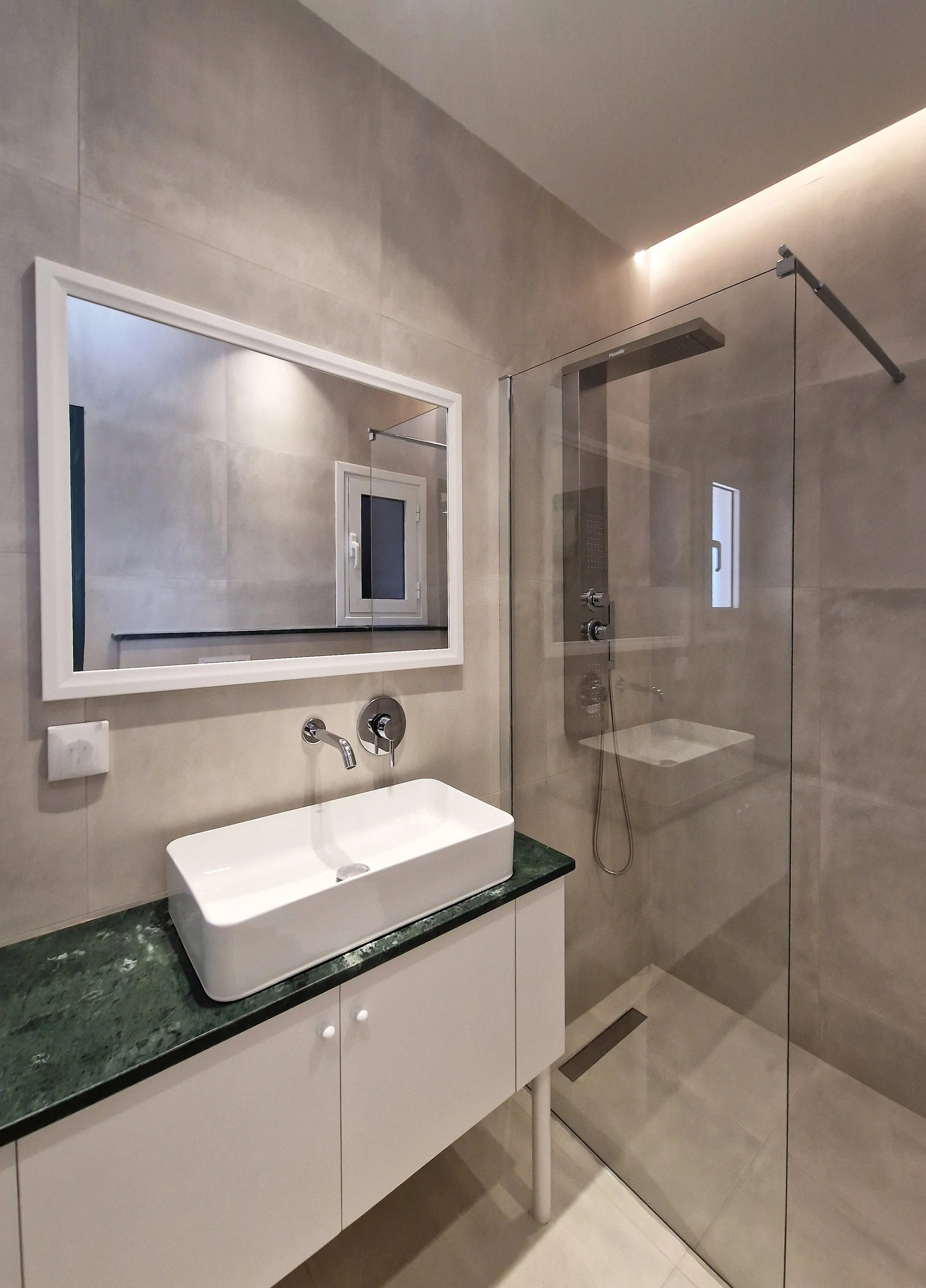 Modern bathroom with gray tile walls, large mirror, white rectangular sink on a green marble countertop, and a walk-in shower with glass partition and rain shower head.