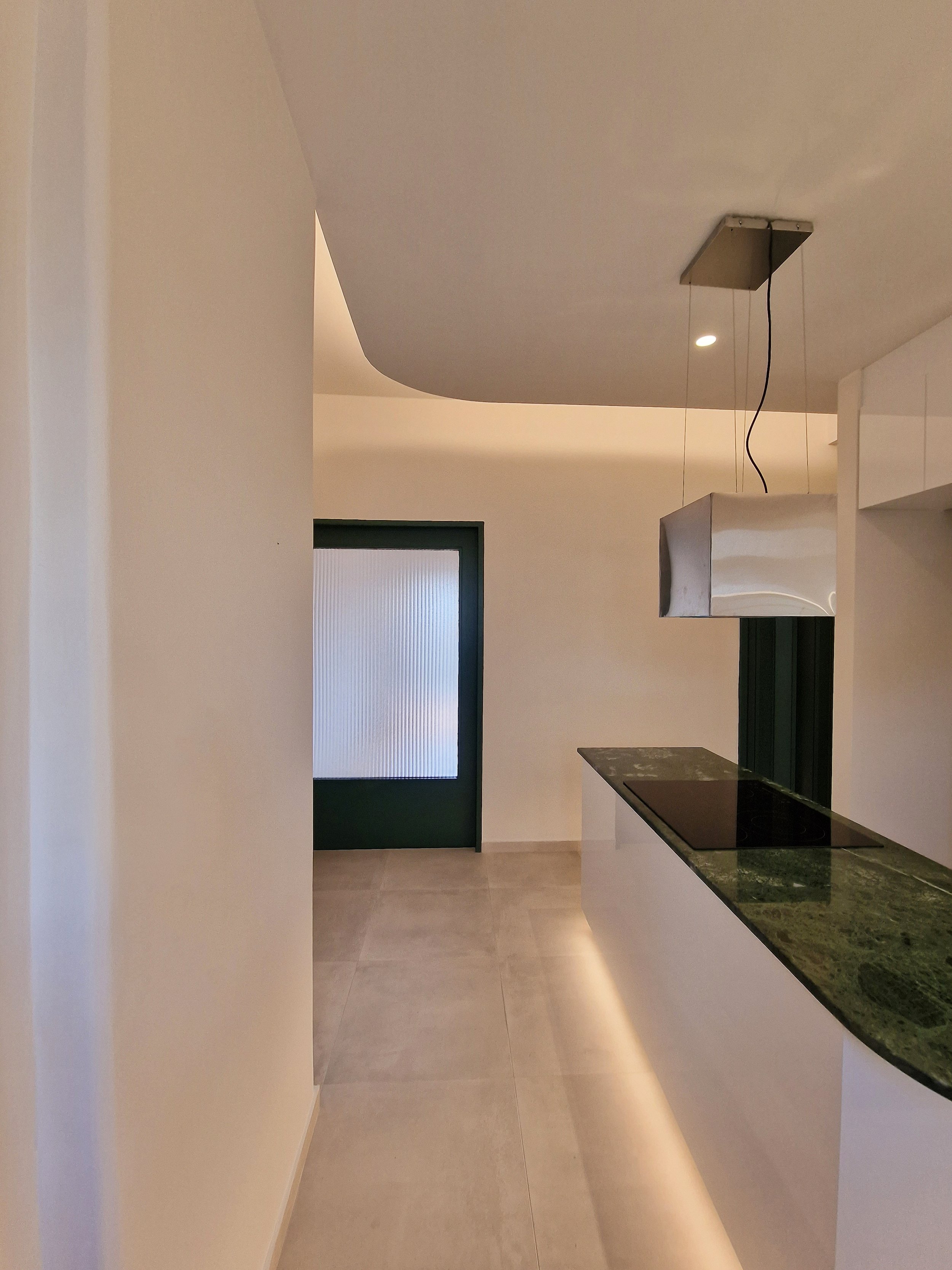 Interior of a modern kitchen with a dark green door, beige tiled floor, white walls, and a countertop with a black cooktop and green marble surface.