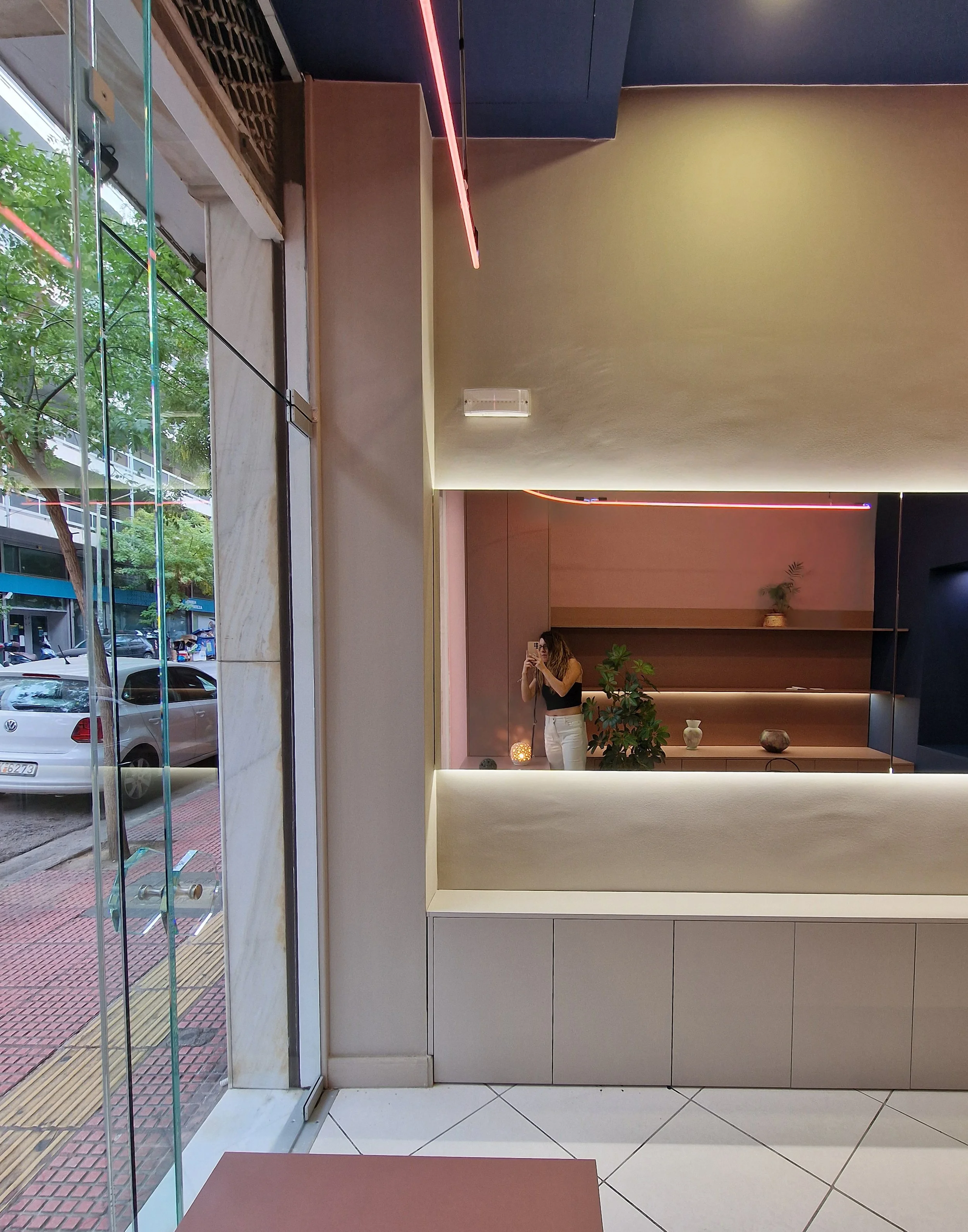 Interior of a modern, minimalistic building lobby with a large mirror, potted plants, decorative vases, and a woman taking a photo. Outside, cars and trees are visible through large glass windows.