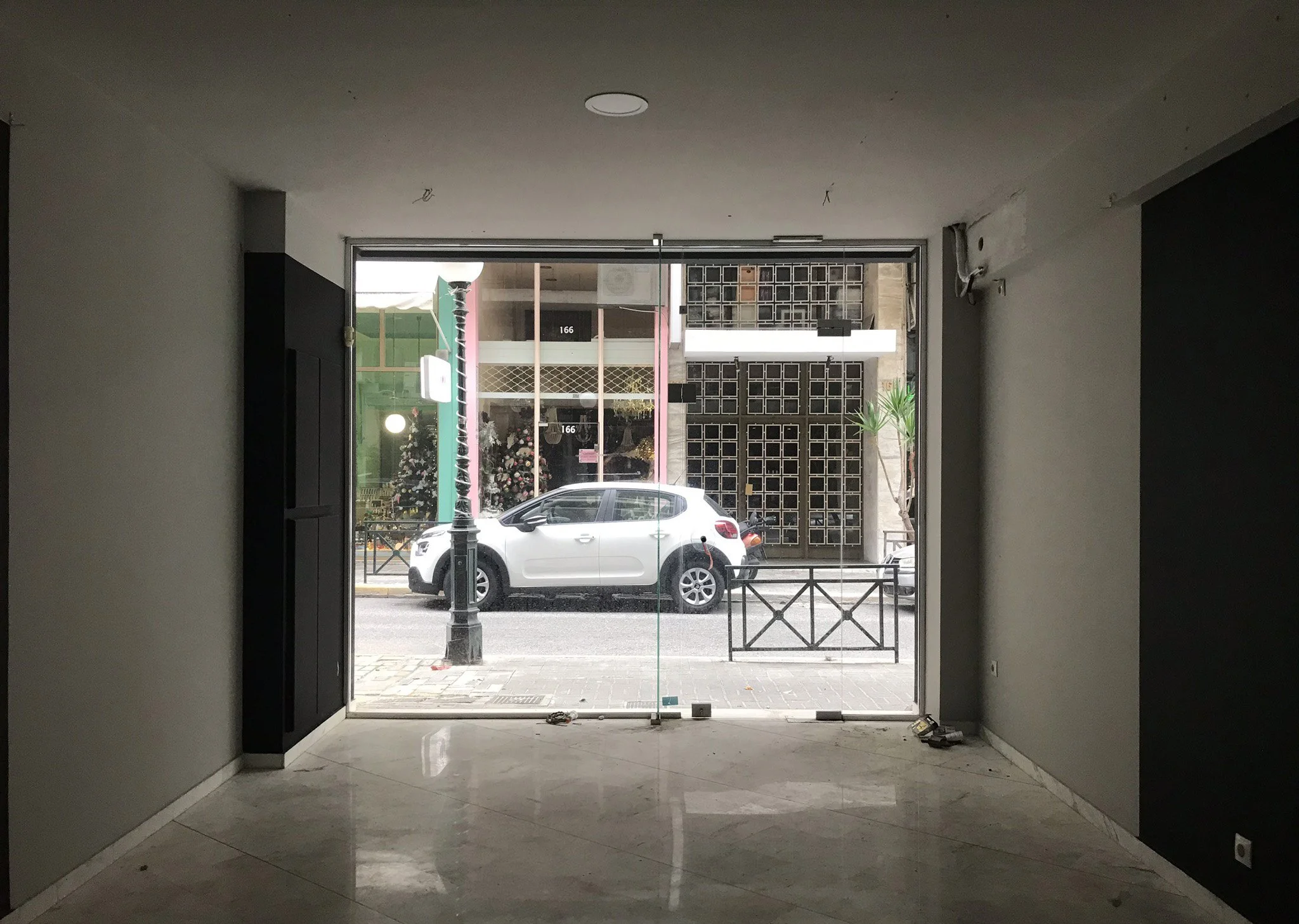 Interior view of an unfinished building with a glass door leading to a sidewalk and street outside, including a parked white car, streetlamp, and a decorated Christmas tree visible through the glass.
