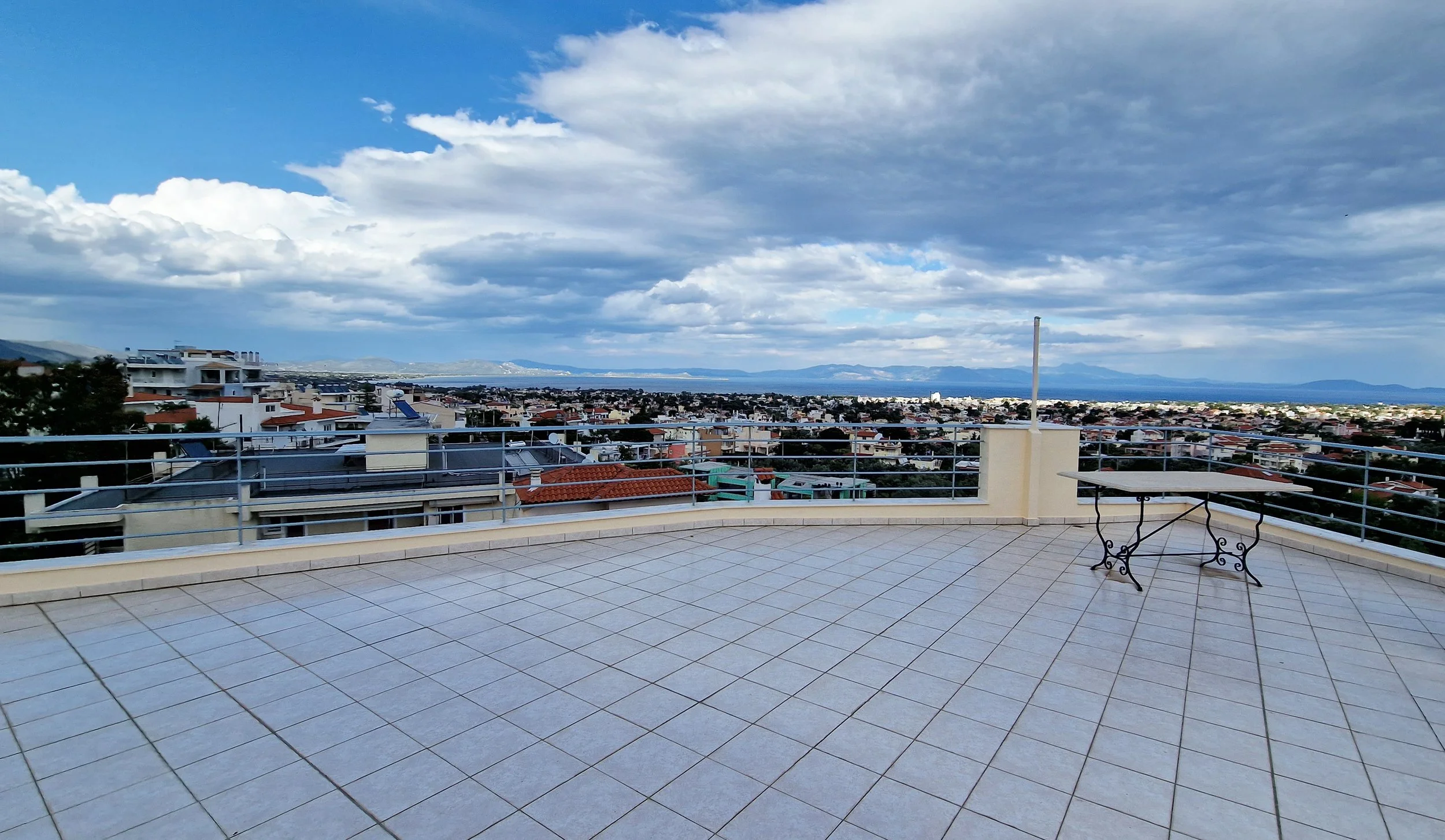 View from a rooftop terrace with tiled flooring, metal railing, a small table with ornate metal legs, overlooking a cityscape with white buildings, red-tiled roofs, and a distant water body under a partly cloudy sky.