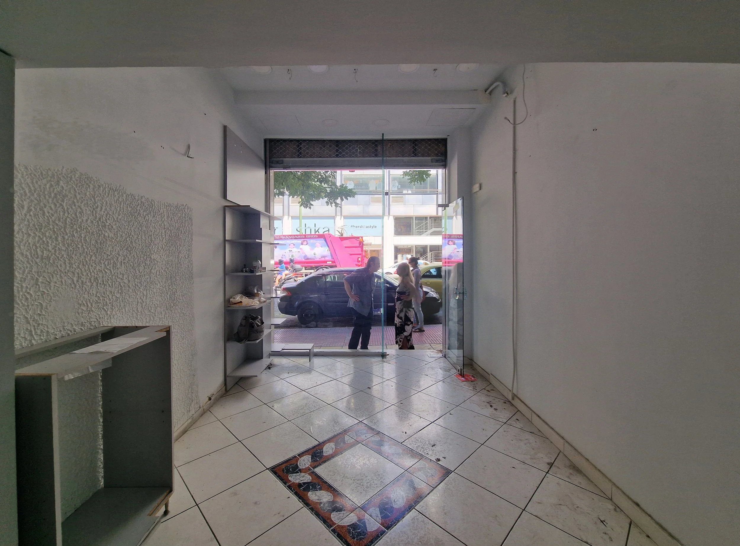 Interior view of a storefront with an open glass door, showing parked cars, people walking outside, and buildings across the street.