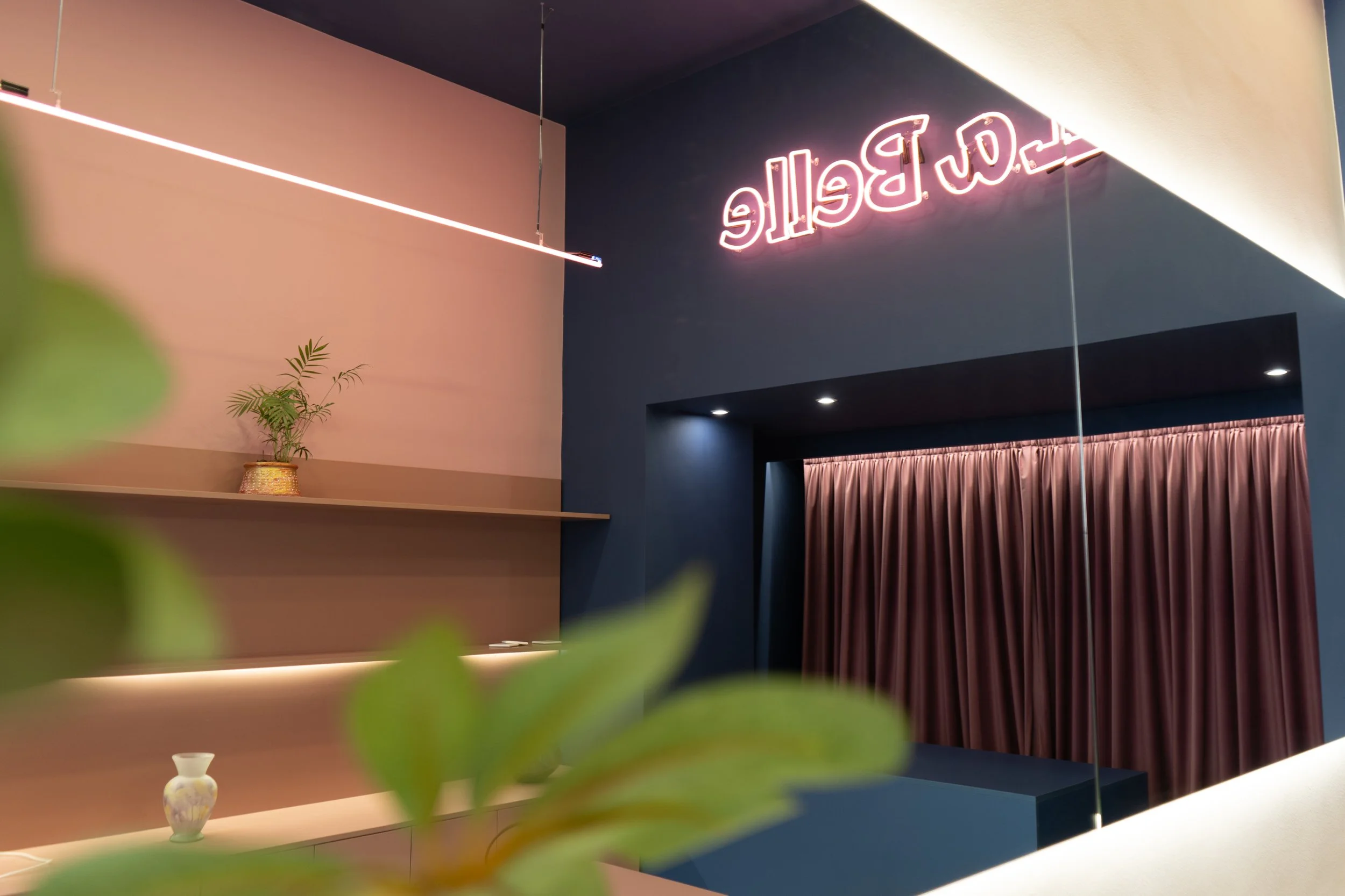 Interior of a modern space with a pink and dark blue wall, a pink neon sign, a shelf with a potted plant and a small decorative vase, and pink curtains.