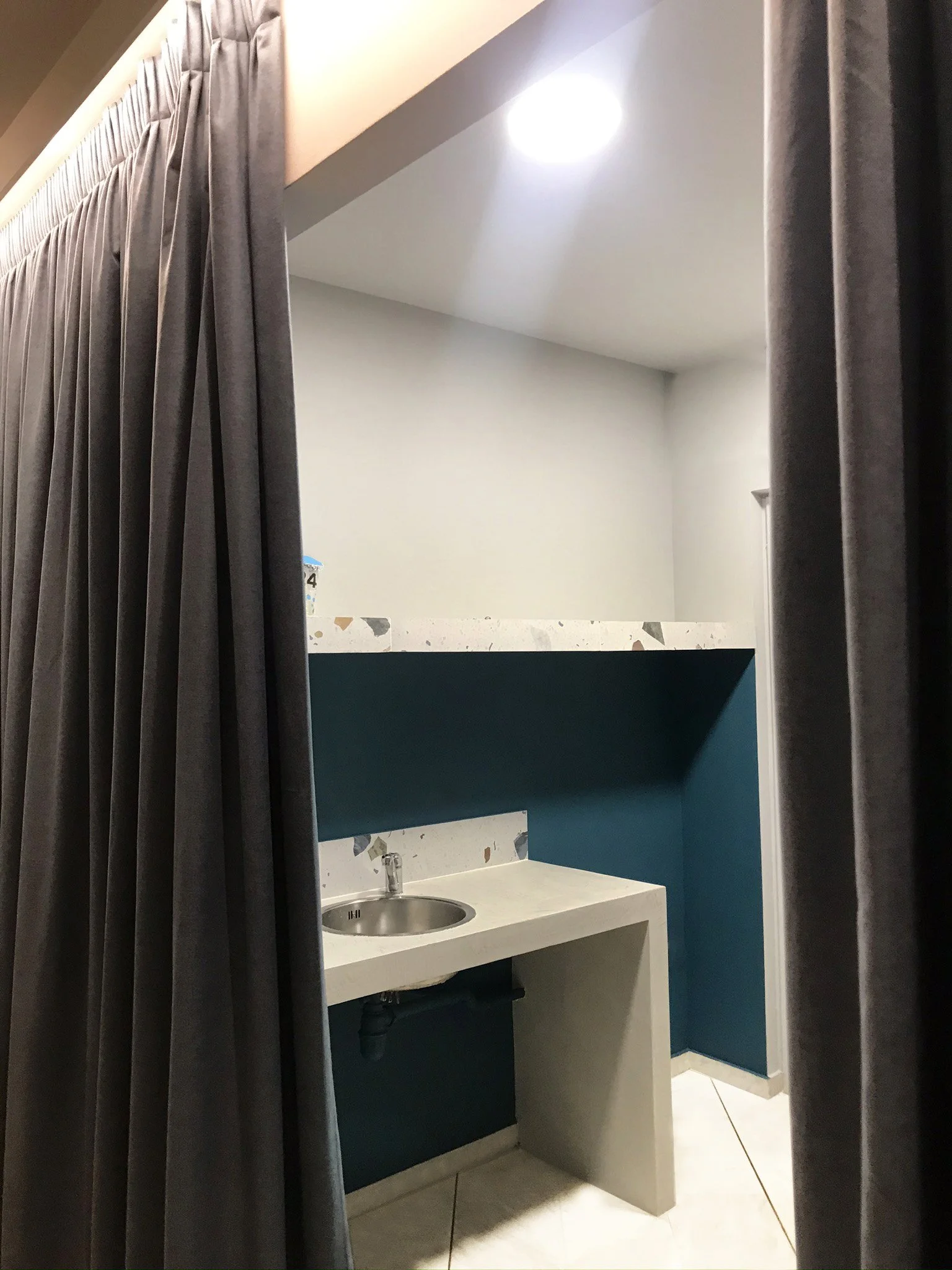 A small bathroom with a white sink, a silver faucet, a dark teal cabinet below, and a decorative backsplash with mosaic tiles. The bathroom has gray curtains on the sides and a light-colored tiled floor.