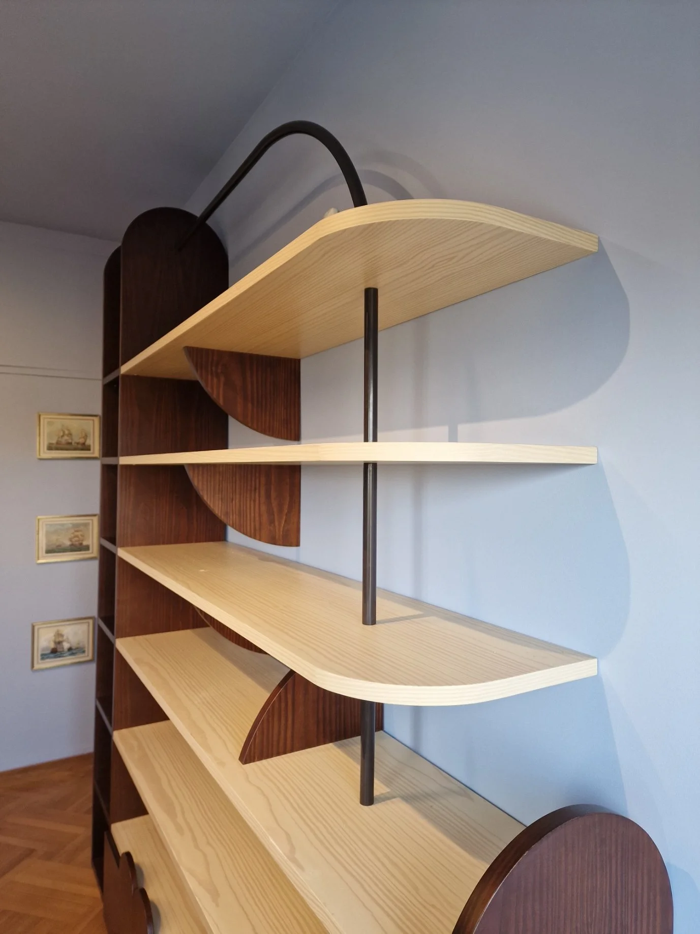 Image of a multi-level wooden bookshelf with curved edges, light-colored shelves, and dark brown vertical supports, set against a blue wall with small framed pictures.
