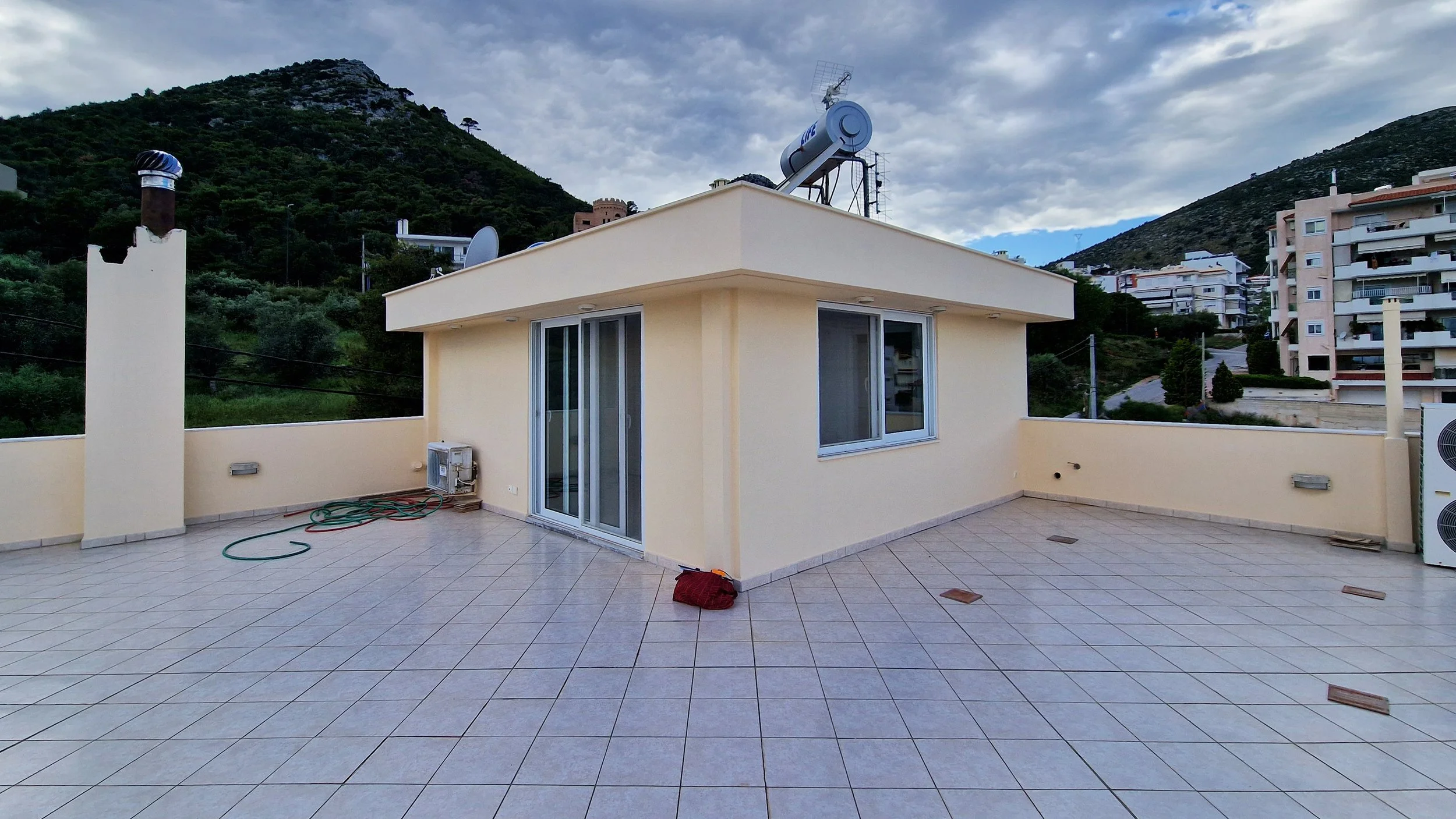 The image shows a rooftop terrace with beige tiled flooring, a beige building with sliding glass doors, and a view of a hillside with other apartment buildings and green vegetation under a partly cloudy sky.