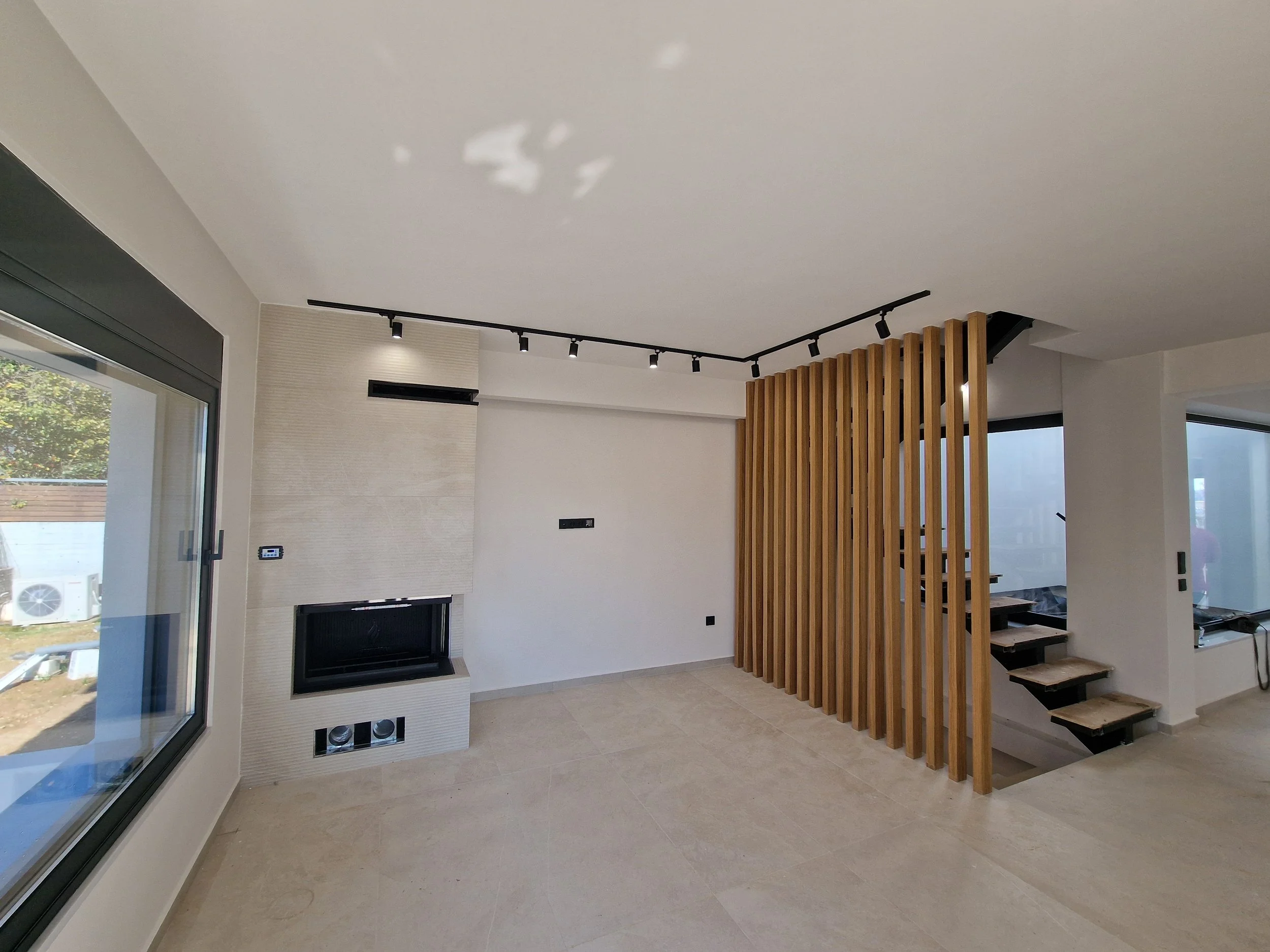 Modern living room with white walls, beige floor tiles, a large glass sliding door on the left, a white fireplace with a textured wall, ceiling track lighting, a wooden slat room divider, and staircase with black steps on the right.