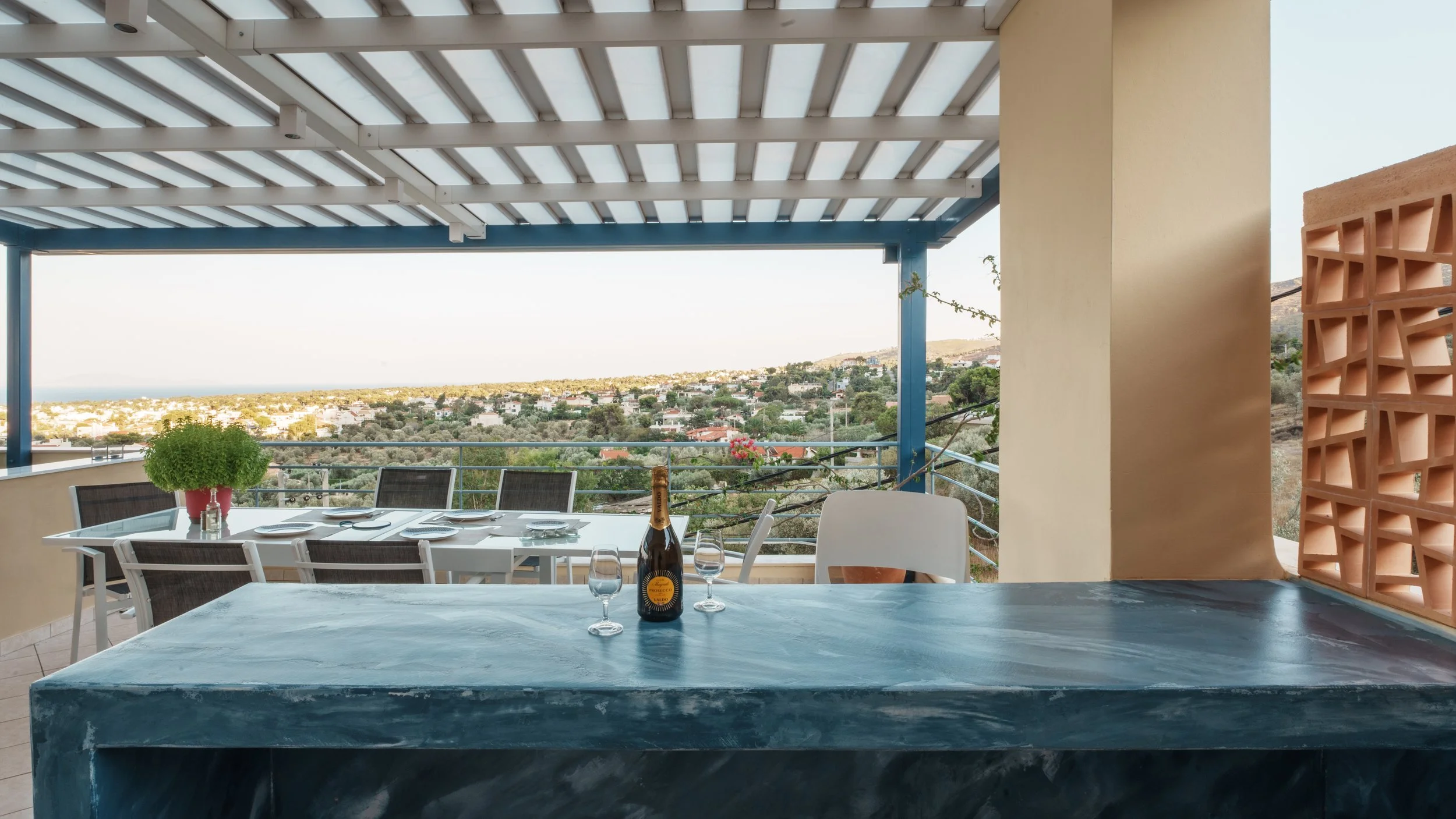Balcony dining area with a table set for four, a bottle of sparkling wine, and a view of a hillside town and landscape in the distance.