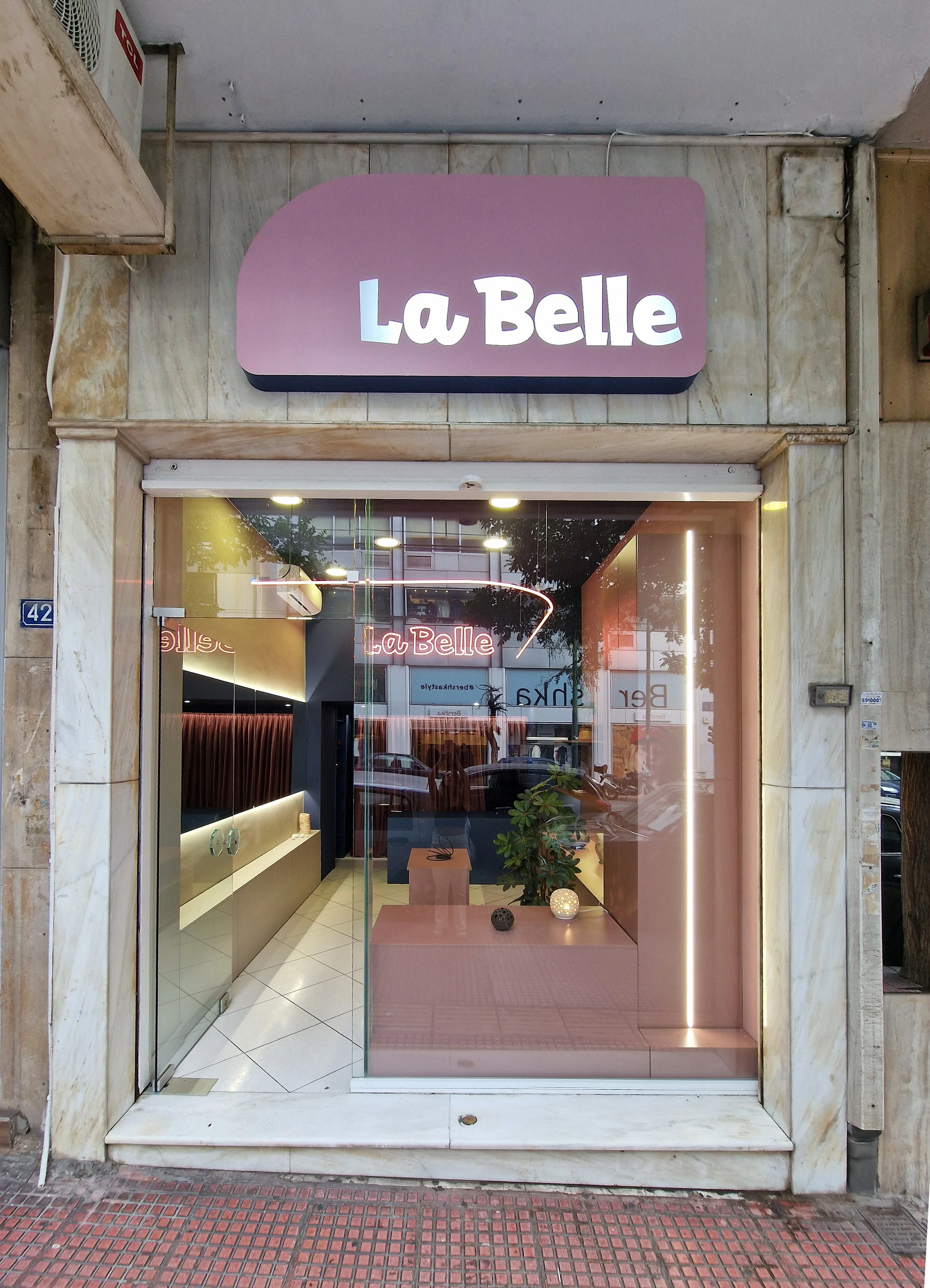 Storefront with a pastel pink and purple sign that says 'La Belle'. Large glass window displaying the interior with a potted plant and decorative items, with a reflection of the street outside. The exterior has a stone facade and a red tiled sidewalk