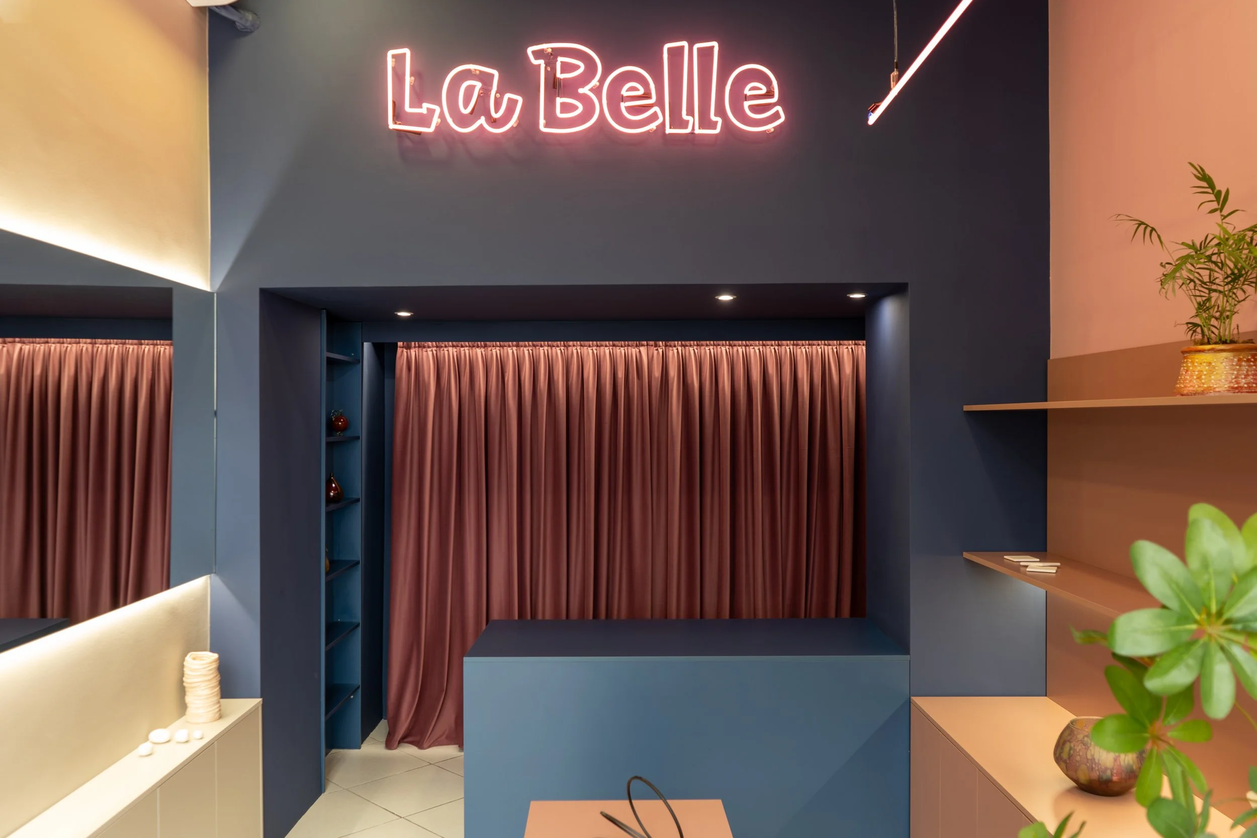 Interior of a modern cafe with a neon sign that says 'La Belle'. The space features a blue and beige color scheme with pink curtains, decorative vases, and a potted plant on a beige shelf.