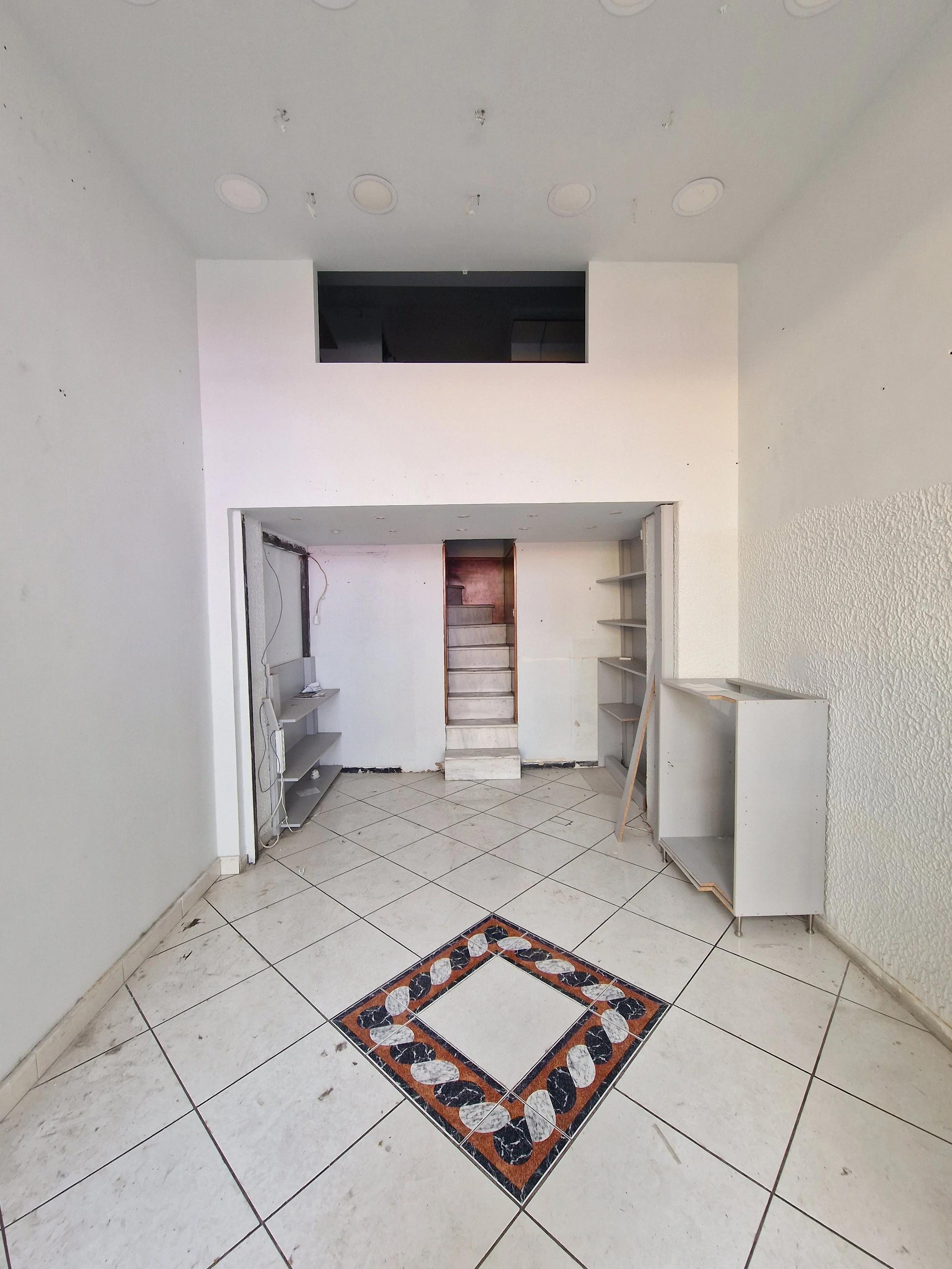 An unfinished room with white walls and decorative tiled flooring. There are built-in shelves on the right wall and a staircase in the back. The ceiling has several circular cutouts and there is a rectangular opening in the wall above the staircase.