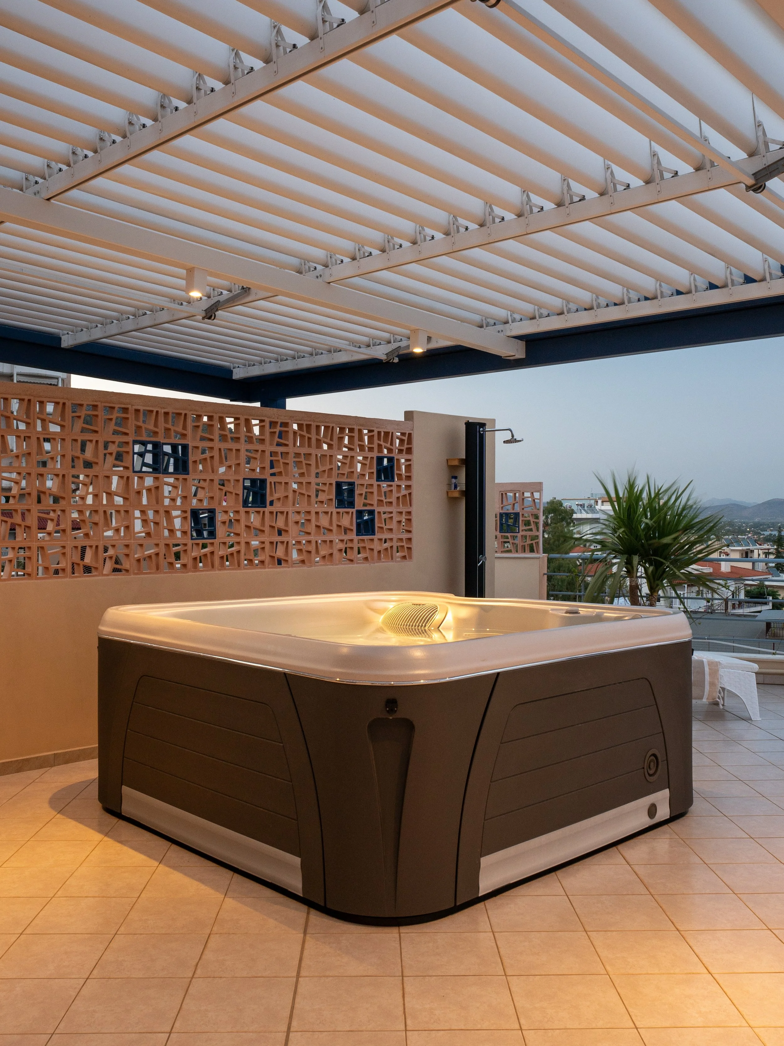 An outdoor rooftop area featuring a hot tub hot tub with beige tiles, a decorative brick wall with small windows, a shed with an outdoor shower, and a view of distant mountains under a partly cloudy sky.