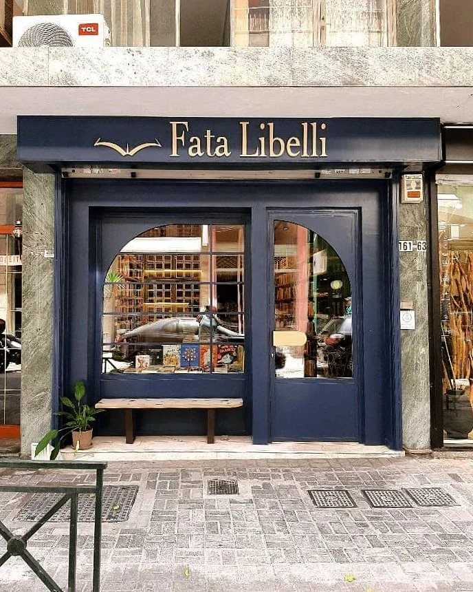 Storefront with a black awning labeled "Fata Libelli" in gold lettering, and a large front window with a bench outside. Potted plant to the left of the entrance.
