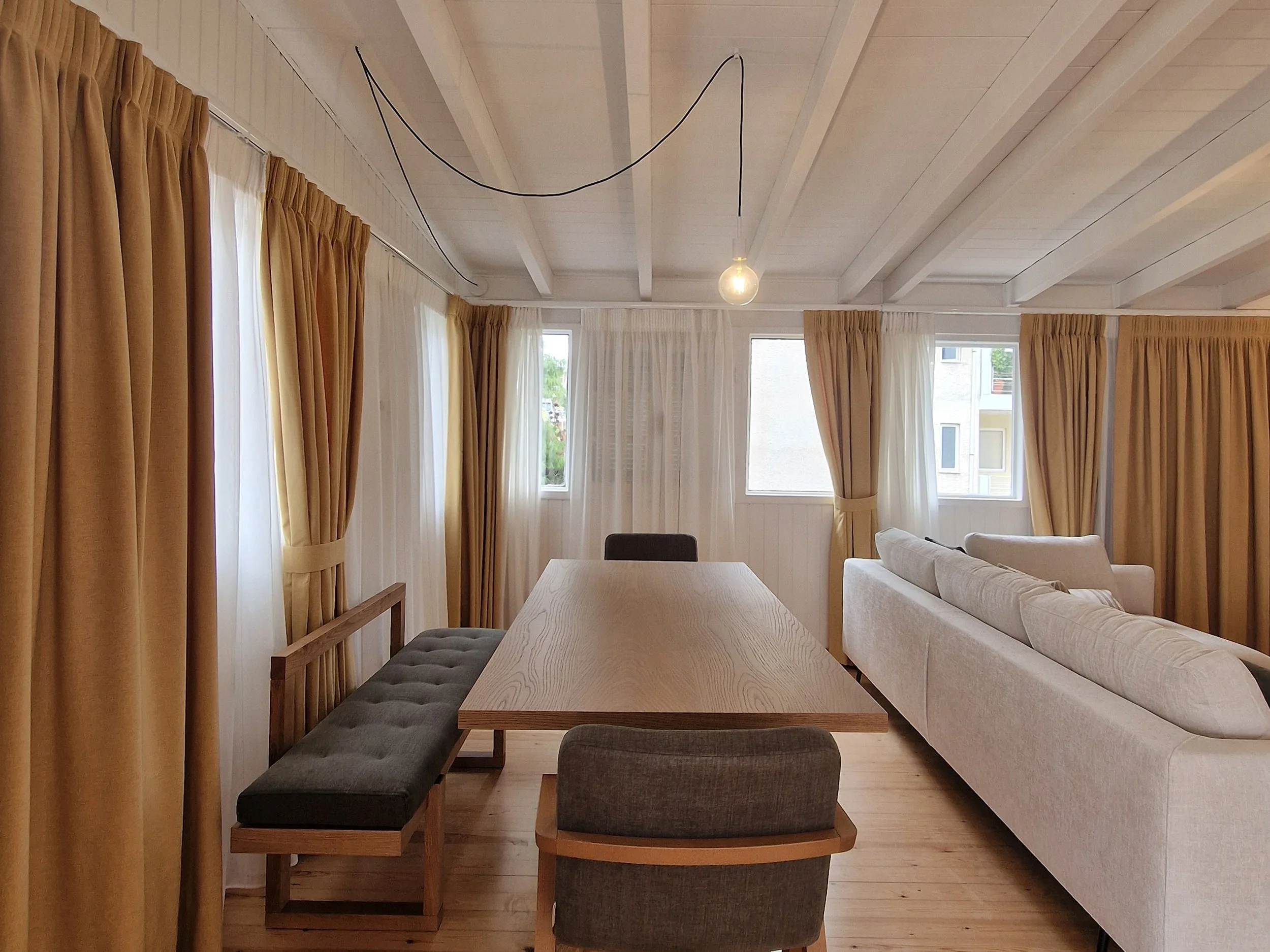 Interior of a living room with beige curtains, large windows, a white sofa, a wooden dining table, and gray cushioned chairs, with a wooden floor and a ceiling light bulb hanging from black cords.