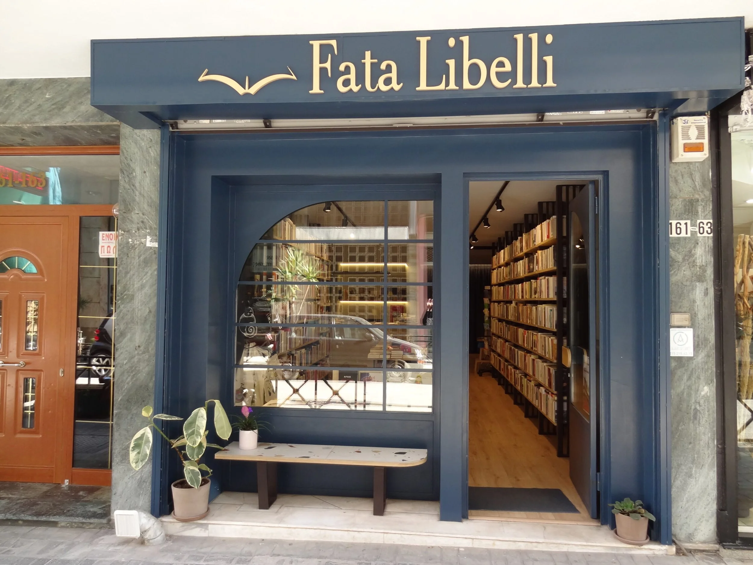 The entrance to a bookstore named Fata Libelli with a dark blue facade, large window showing bookshelves inside, potted plants outside, and a wooden door to the left.