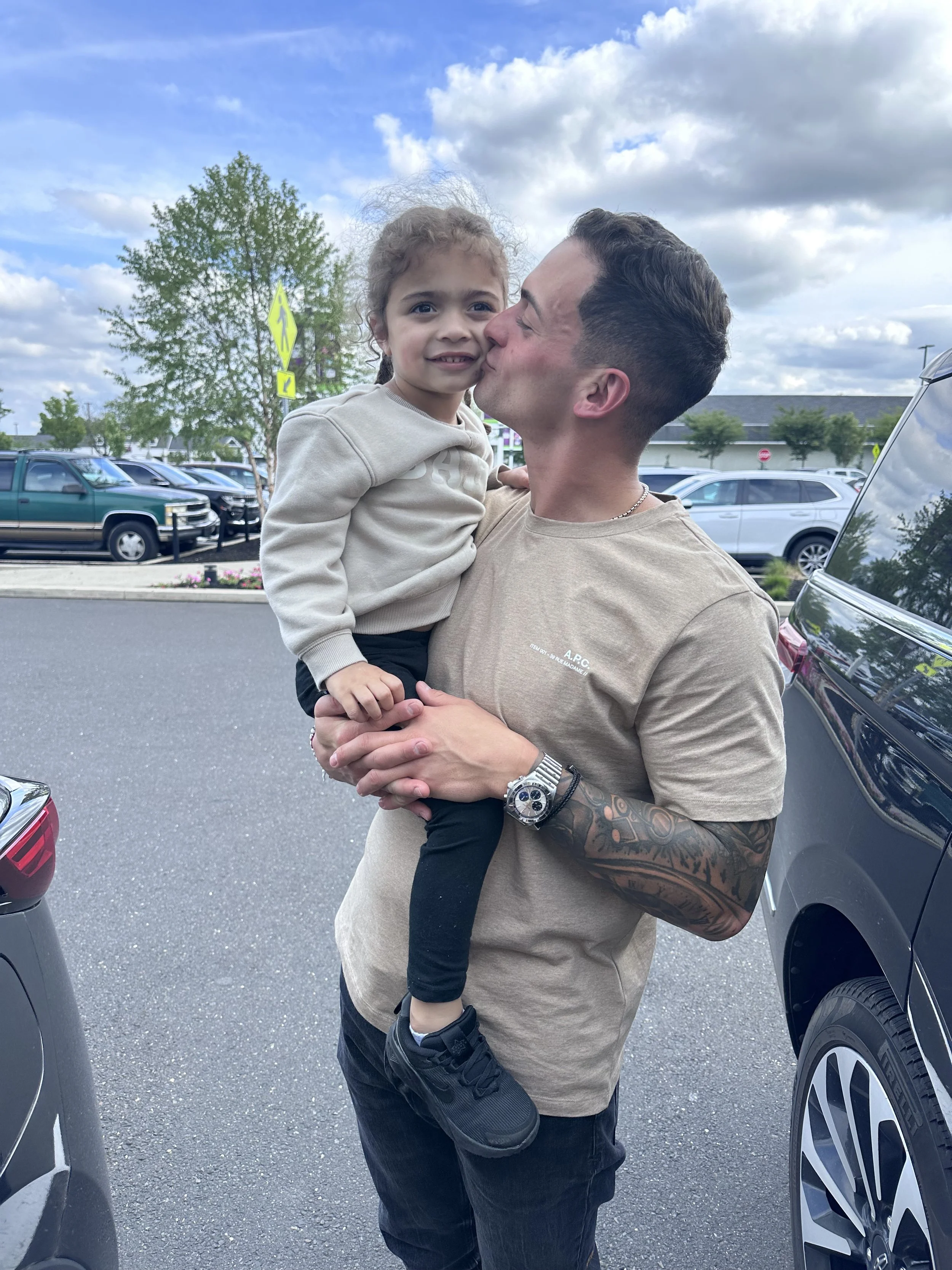 A man holding a young girl outdoors in a parking lot, with a cloudy sky and trees in the background. The man is kissing the girl on the cheek while she smiles at the camera.