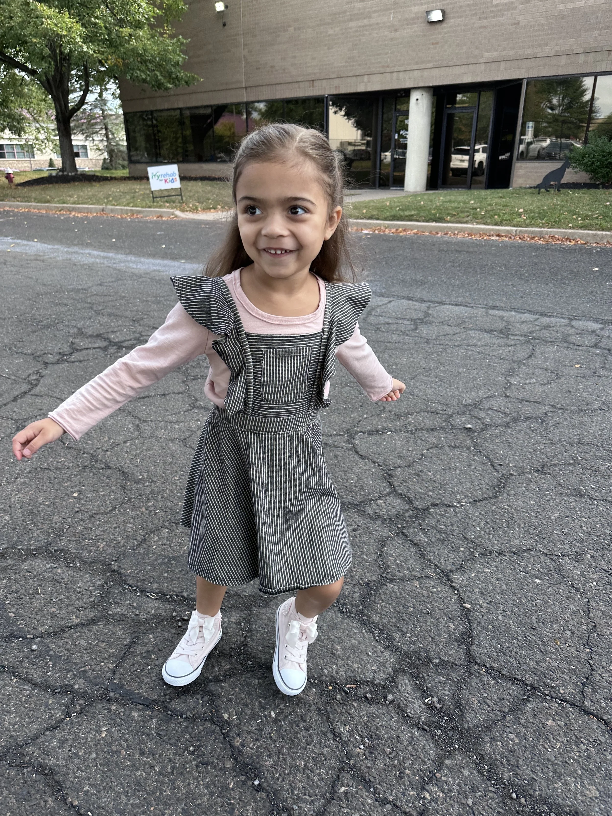 A young girl with long hair smiling and playing outside on paved ground near a building with large windows. She is wearing a striped dress with ruffled shoulders, a long-sleeve pink shirt underneath, and pink sneakers.