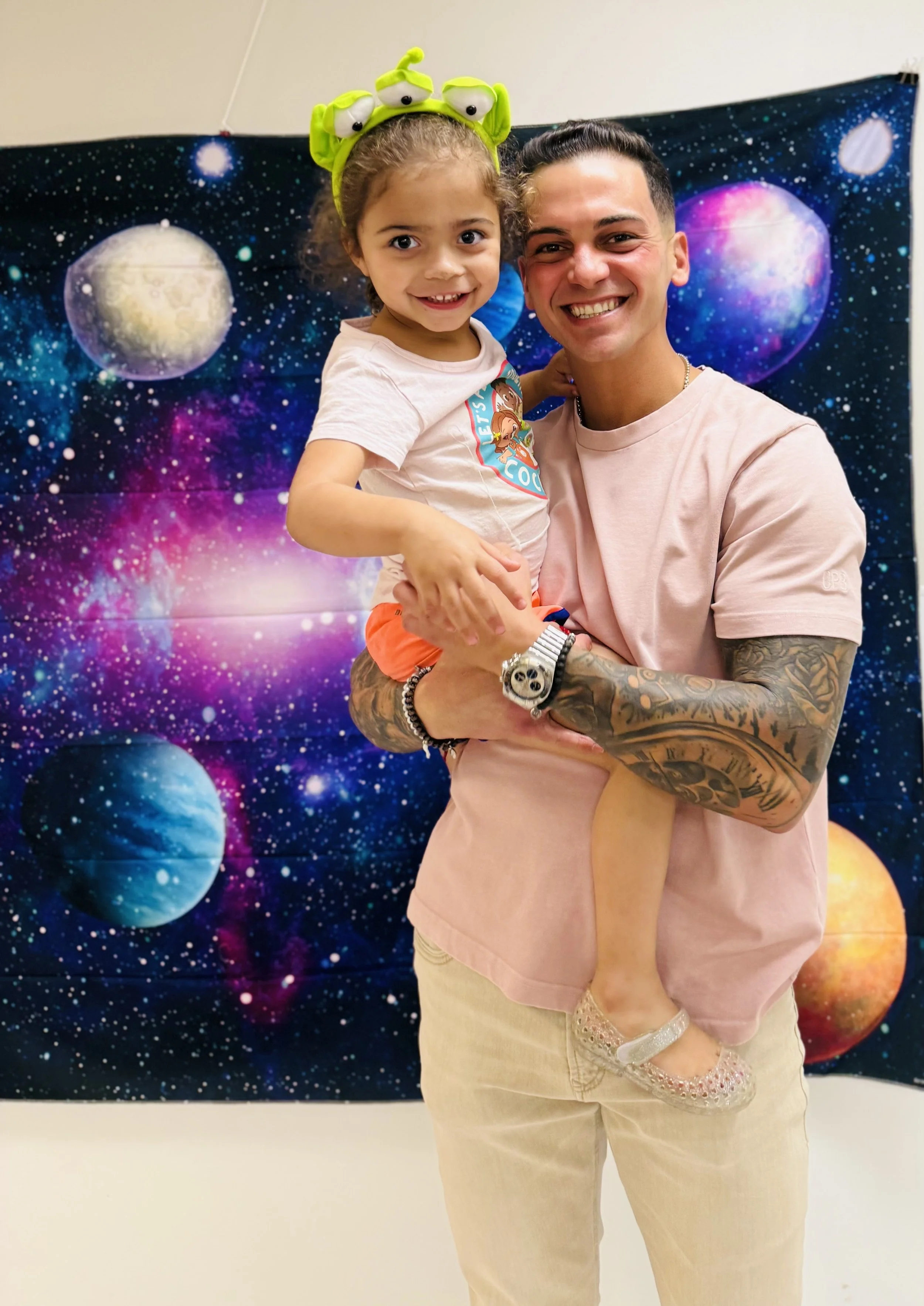 A man with tattoos holding a young girl in front of a space-themed backdrop with planets and stars. The girl is wearing a headband with green alien eyes and a white shirt.