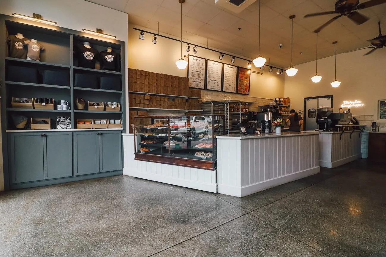 Interior of a small coffee shop with a display case of pastries, a counter with a coffee machine, menu boards on the wall, and a storage shelf with merchandise. The shop has hanging lights and ceiling fans.