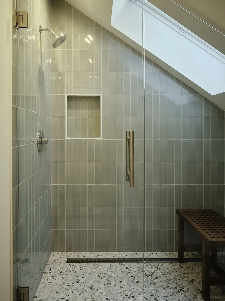 Modern walk-in shower with tiled walls and a skylight, featuring a wooden bench and a built-in niche for toiletries.