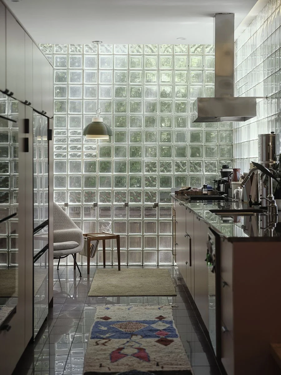 A modern kitchen with glass block wall, pendant light, and countertop appliances, featuring a small sitting area with a chair, a side table, and colorful rug.