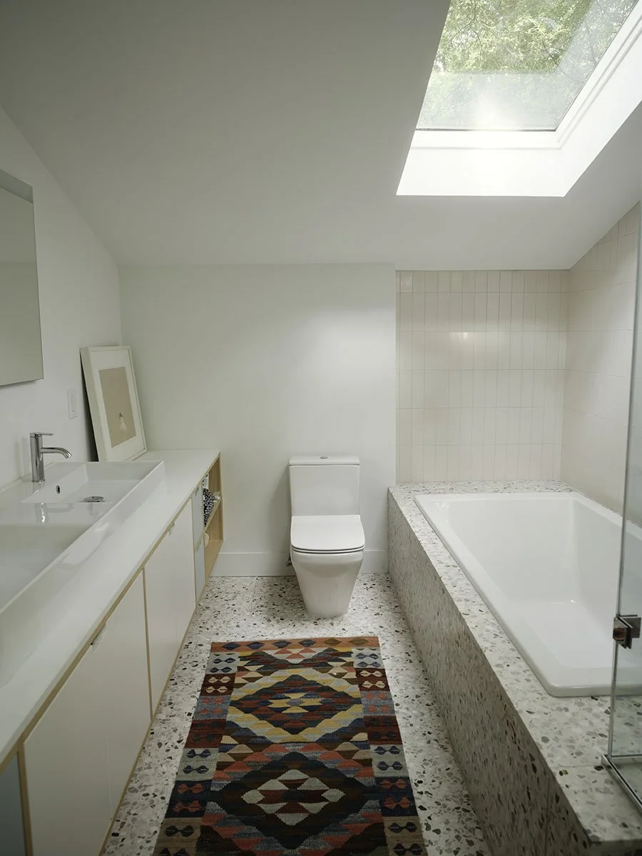 A modern bathroom with a skylight, white walls, a built-in bathtub, a white toilet, a vanity with a sink, a mirror, a colorful patterned rug, and a framed picture leaning against the wall.