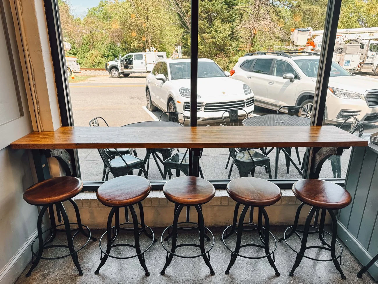 Inside a cafe, a long wooden counter with five wooden stools facing a large window. Outside the window, a parking lot with several white cars and trucks, and trees in the background.