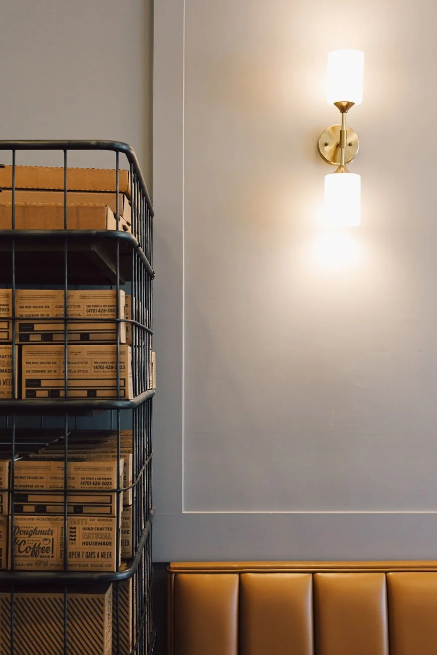 Interior of a cafe or restaurant with a wall-mounted light fixture, a metal wire shelving unit filled with boxes, and a tan cushioned bench.