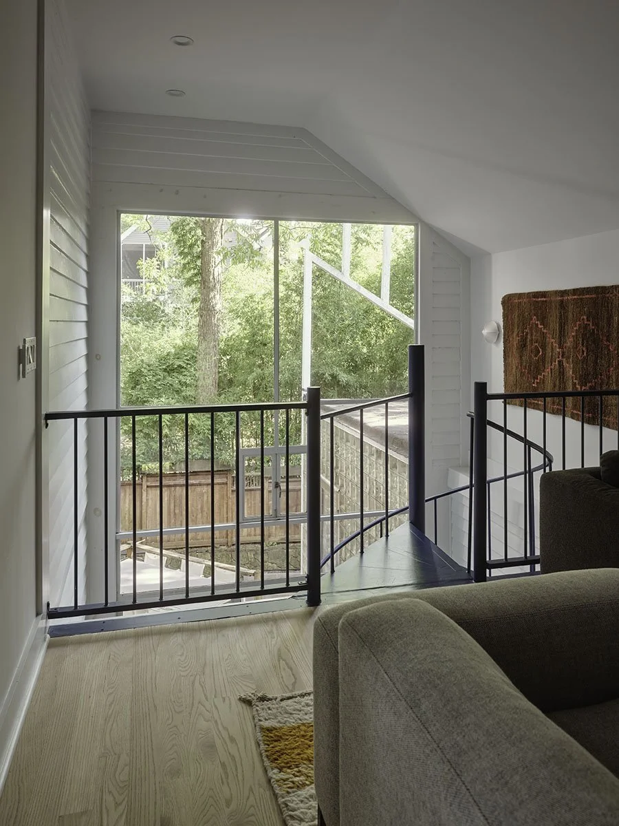 View of the upstairs landing with railing looking out through a large window to trees and a wooden fence outdoors. Part of a beige sofa and a patterned rug are visible inside the room.