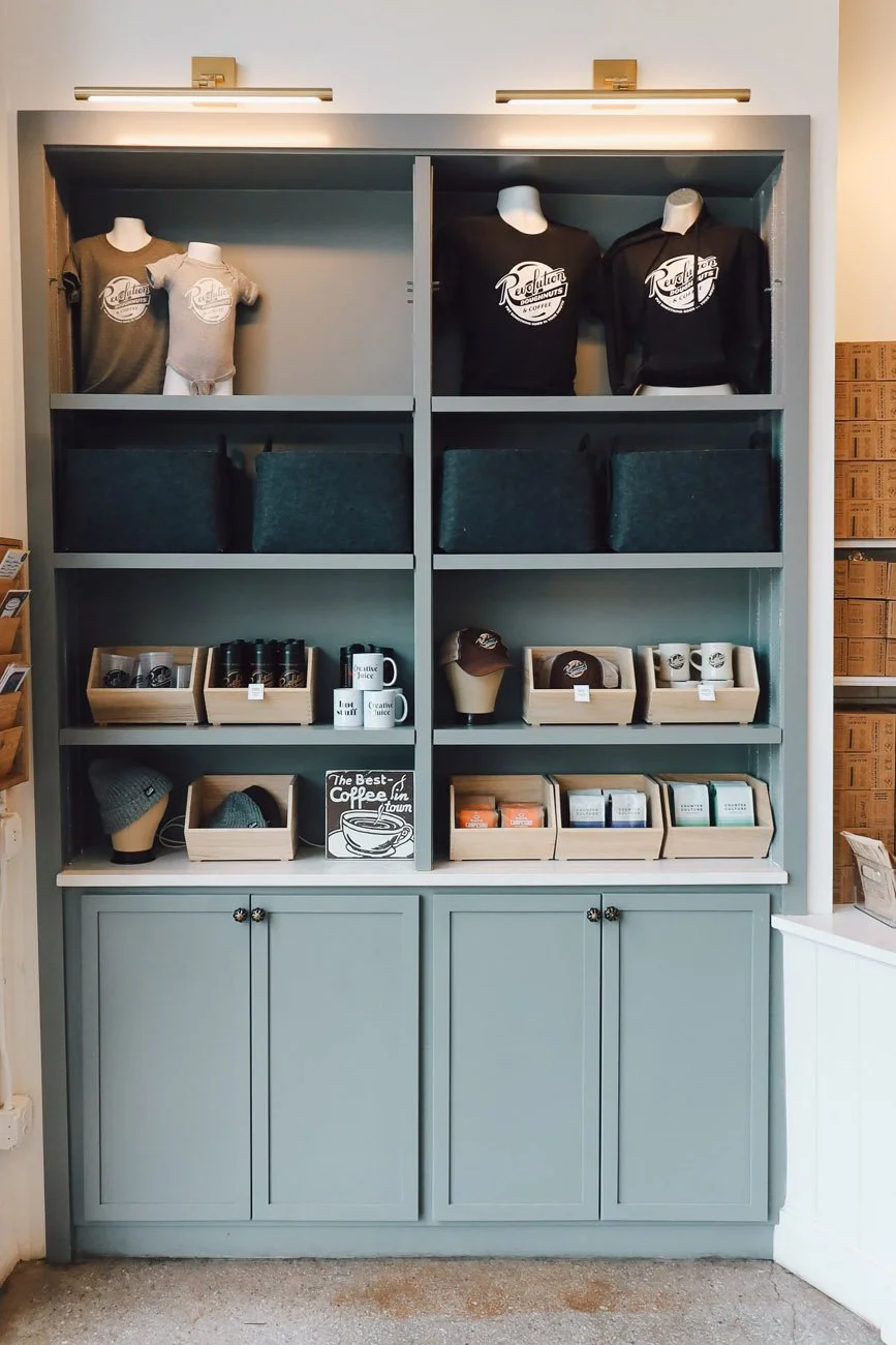 Display shelf with coffee mugs, hats, t-shirts, hoodies, and coffee-related products in a coffee shop.