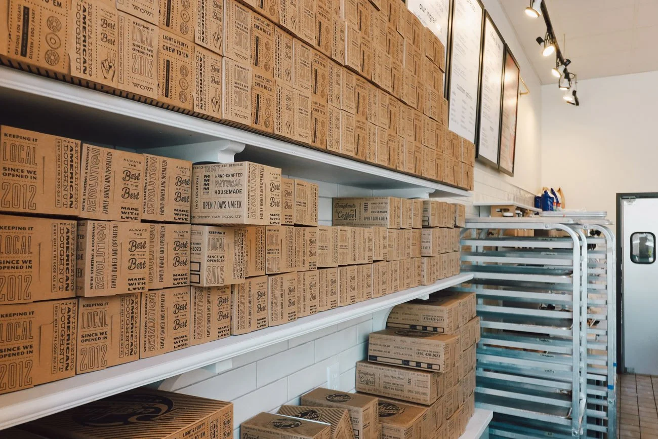 Shelves filled with stacked boxes and cartons of baked goods, possibly pizza or bakery products, in a bakery or cafe setting.