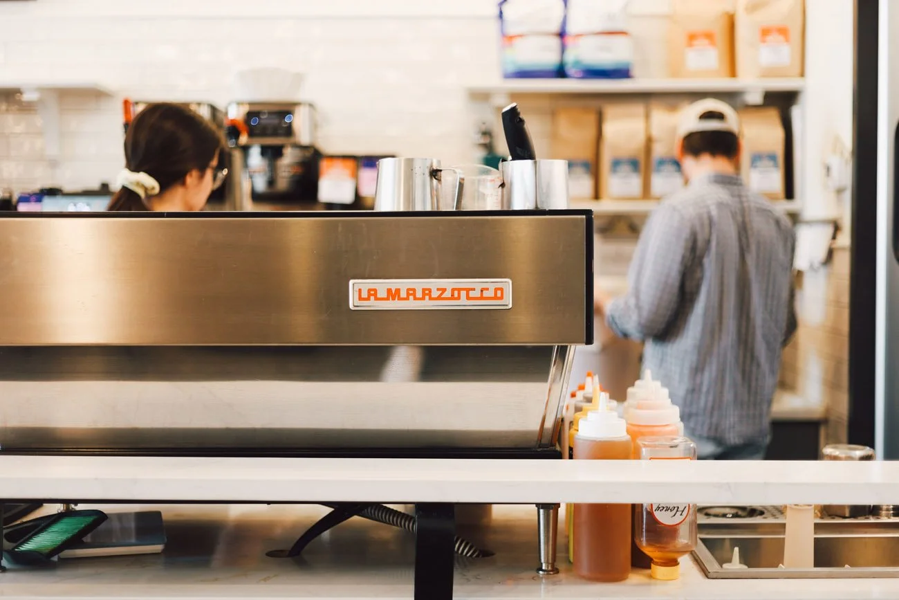 Inside a coffee shop with a stainless steel La Marzocco espresso machine, two employees, shelves with coffee supplies, and condiment bottles on the counter.