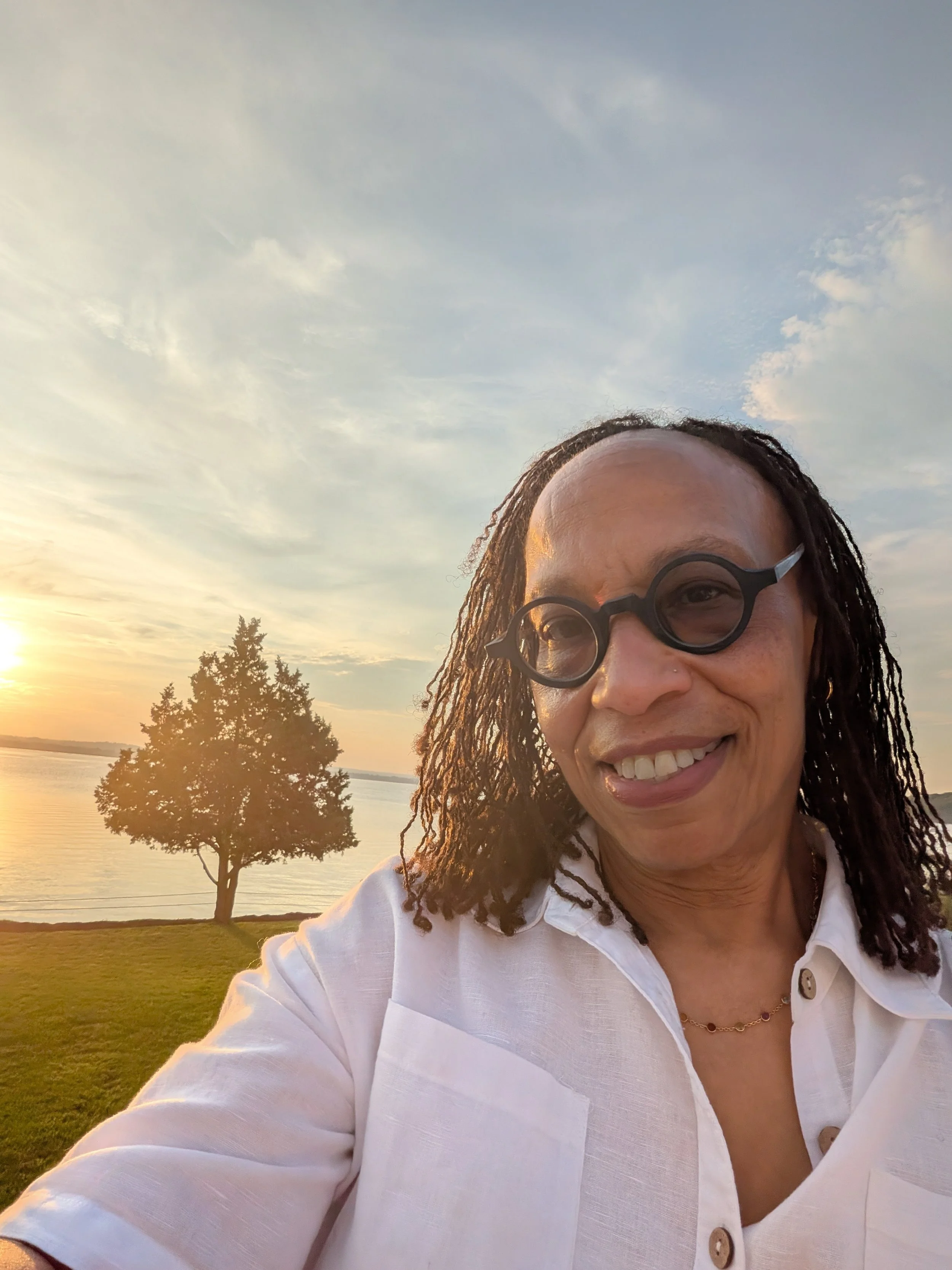 Woman with dark curly hair and glasses smiling outdoors by a lake at sunset, wearing a white shirt.