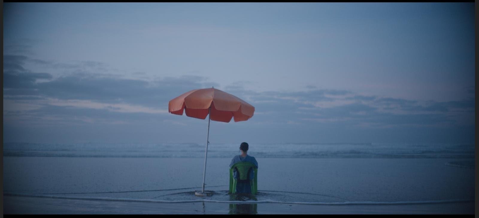 A person sitting in the water on a beach, facing the ocean, under an orange umbrella during dusk or dawn, with cloudy sky.