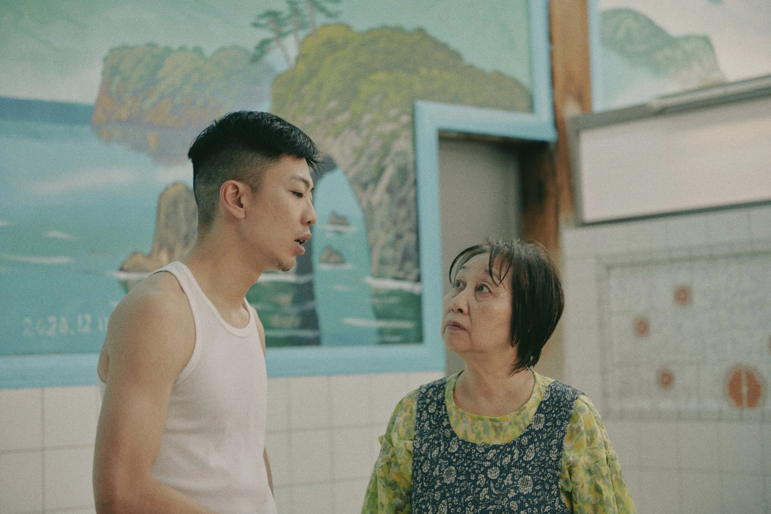 A young man and an older woman talking indoors, with a colorful mural in the background.