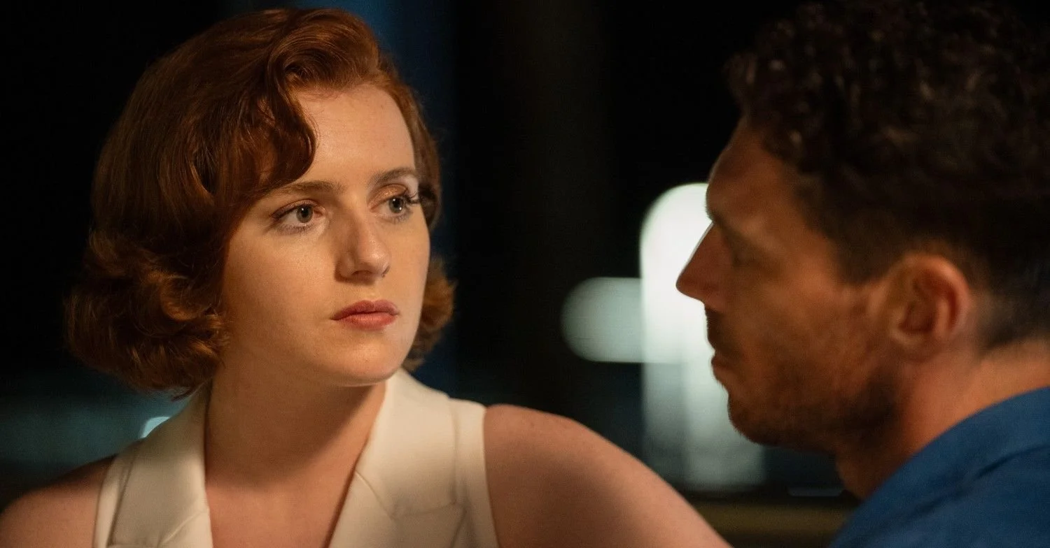 A woman with short red hair and a man with dark curly hair sitting closely during a conversation at night with blurred city lights in the background.