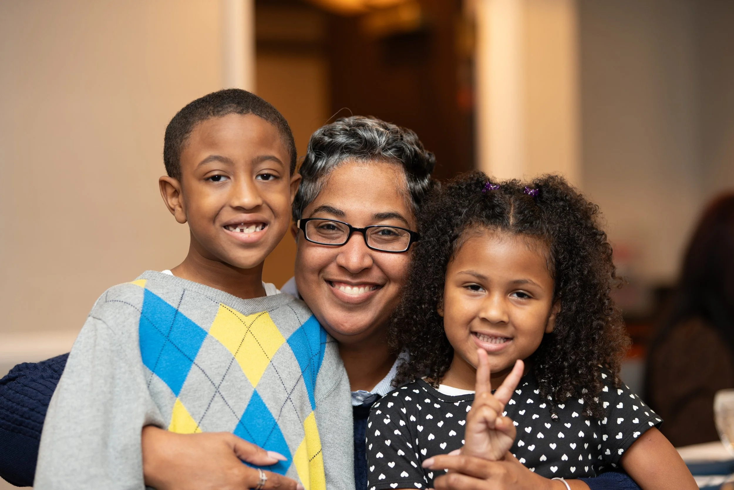 A woman with short gray hair and glasses is hugging two children, a boy and a girl, while smiling at the camera. The boy has short hair and is wearing a colorful sweater, and the girl has curly hair and is wearing a polka-dot shirt.