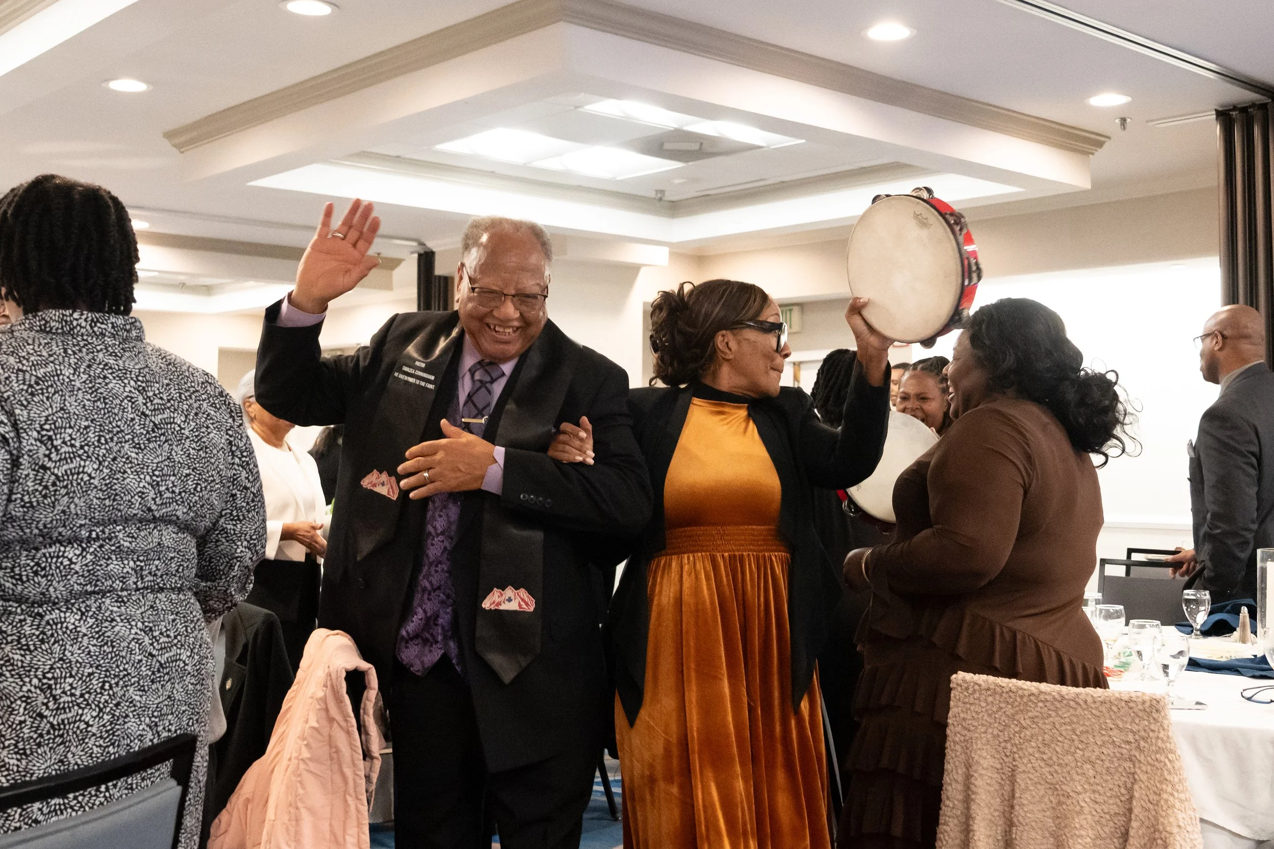 A group of diverse people at a formal event, smiling, laughing, and celebrating. One woman is holding a tambourine, and others are engaged in conversation.