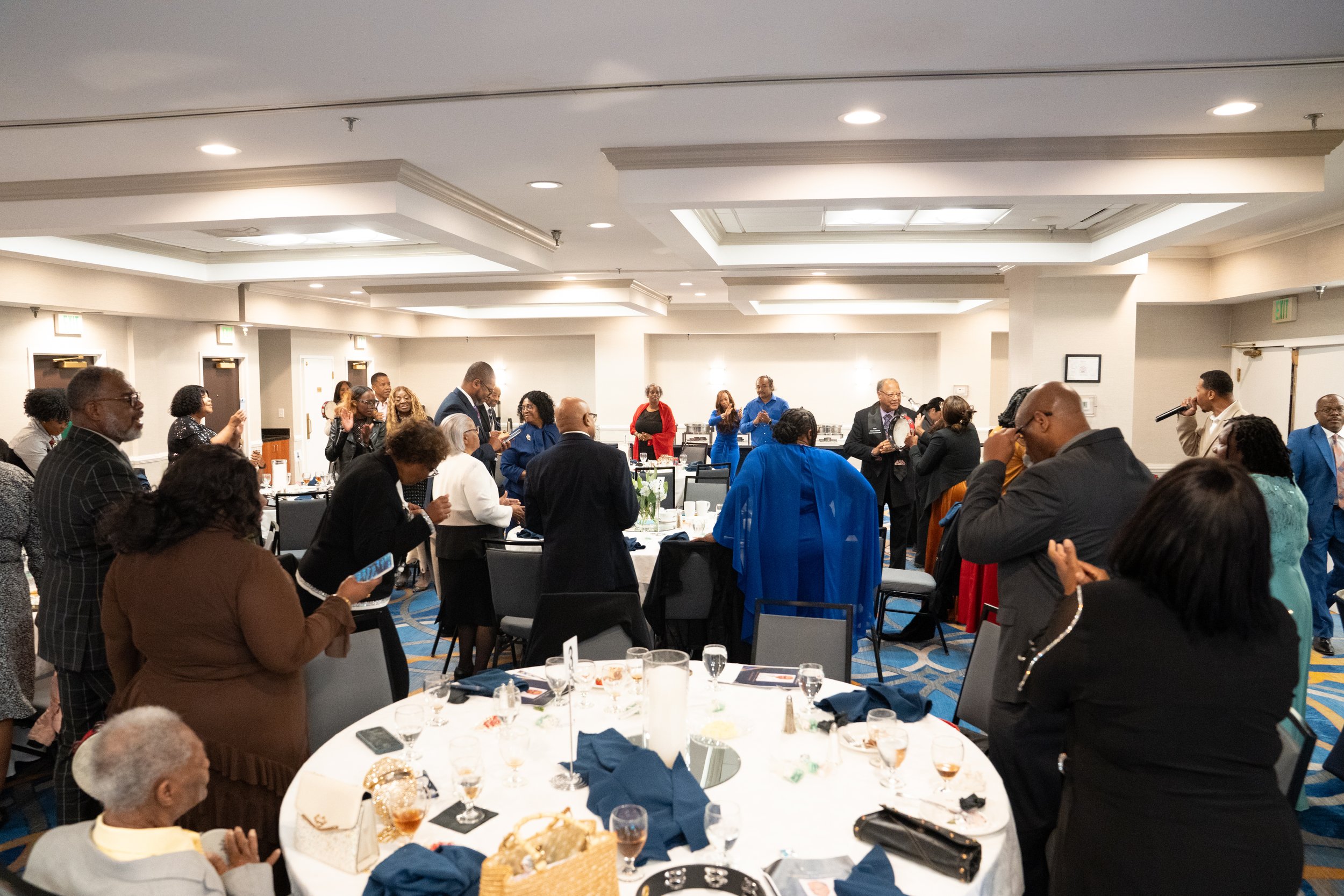 A group of people at a formal event in a banquet hall, some are standing, some sitting at tables with food and drink, and a person is speaking into a microphone.