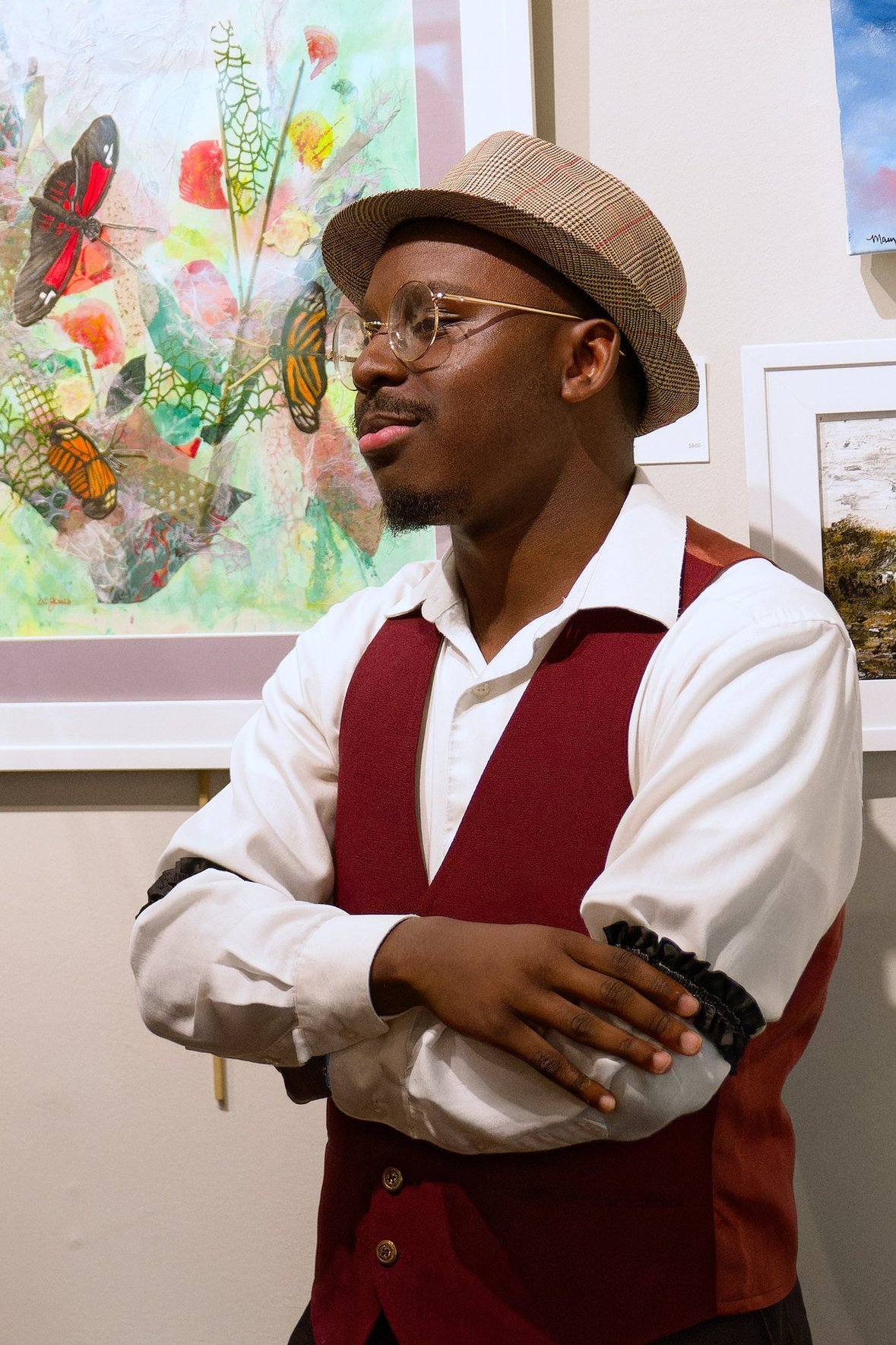 Jalen Collins wearing glasses, a plaid hat, a white shirt, and a red vest with black decorations on the sleeves, standing in front of colorful artwork with butterflies and abstract designs.