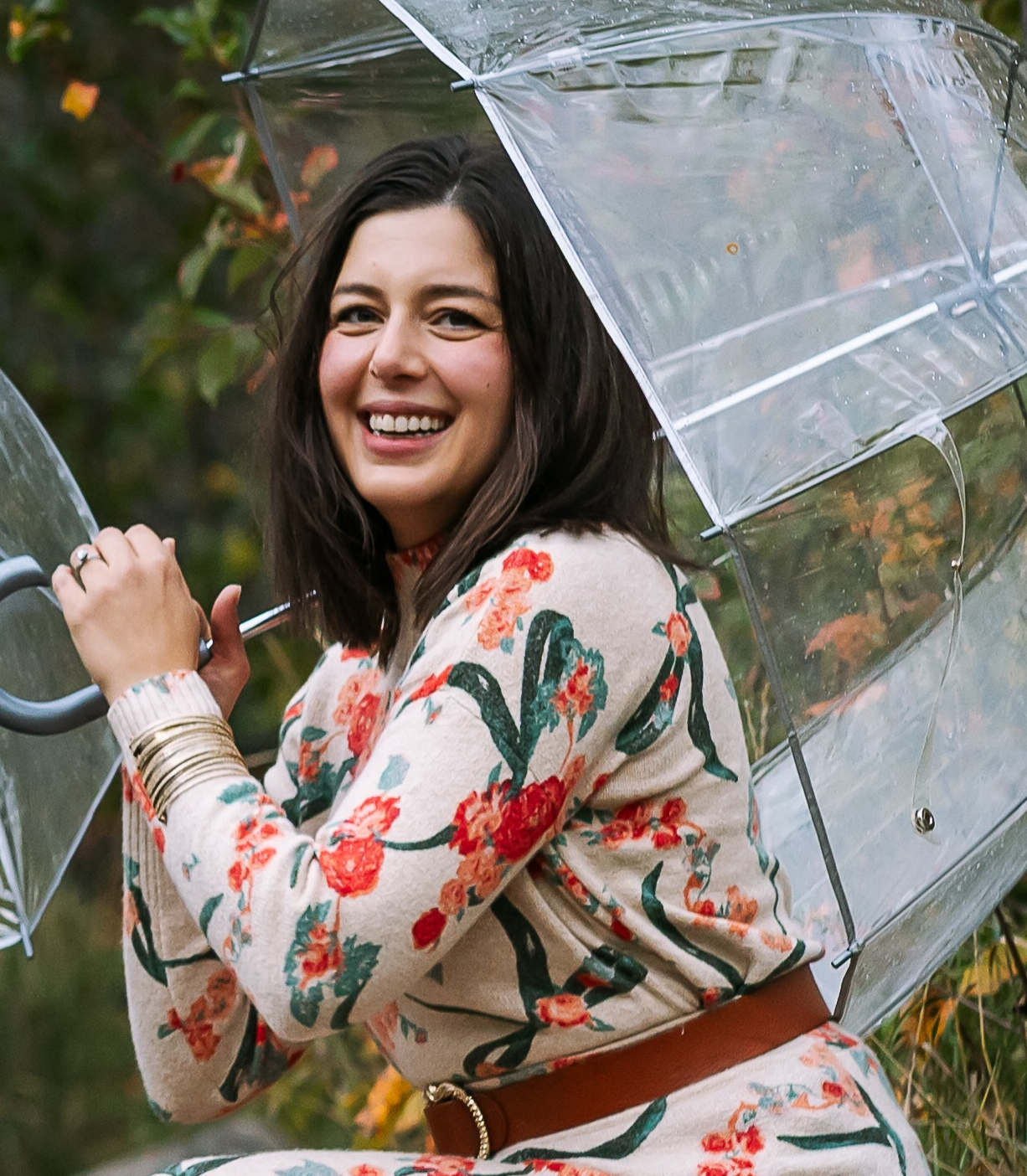 Sara Loca the counselor and coach who owns Forza Tesoro Psychotherapy and Coaching is crouching down with a clear umbrella smiling with fall leaves in the background