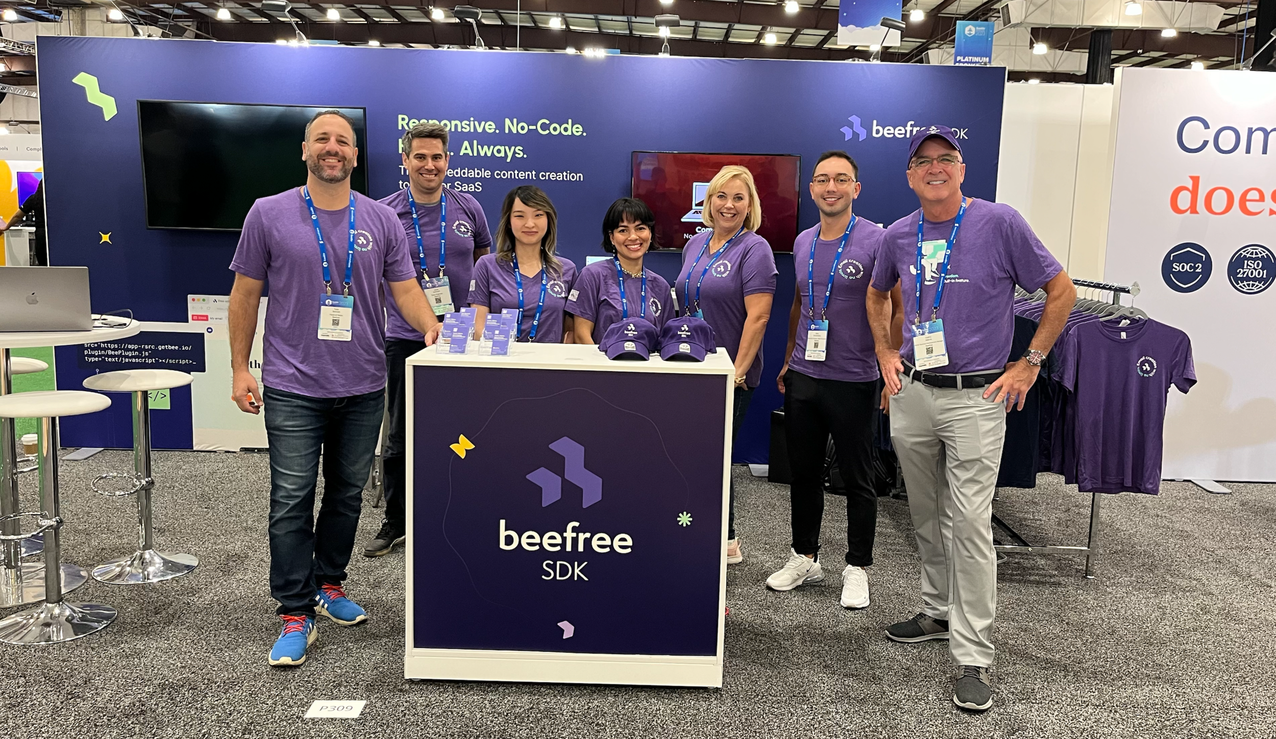 Group of seven people standing behind a booth at a trade show, wearing purple t-shirts with the 'beefree' logo, smiling for the photo. The booth displays a sign with the 'beefree SDK' logo and a screen showing promotional content.