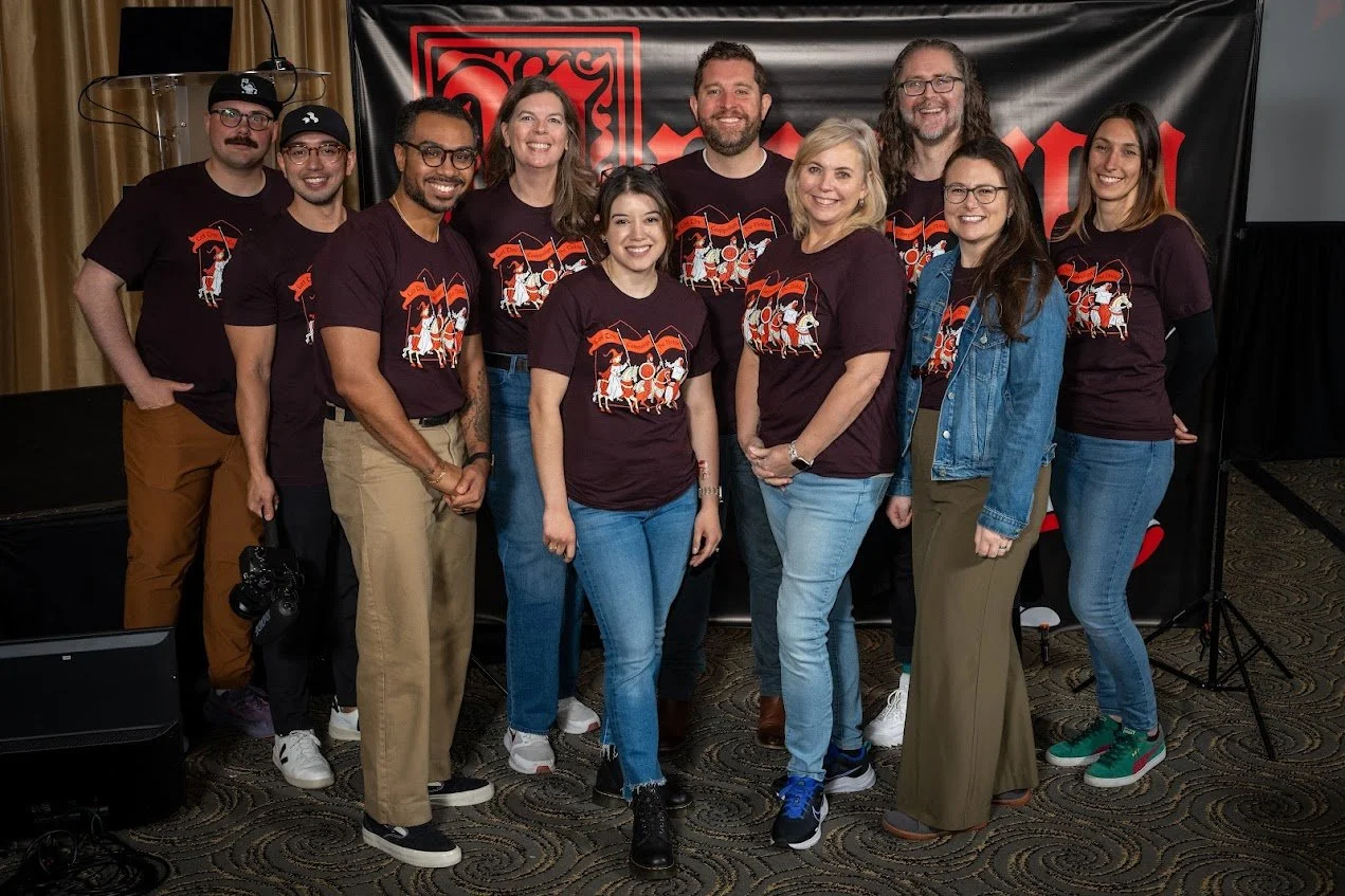 A group of ten diverse people smiling and standing together in front of a black backdrop with red text. They are all wearing matching dark-colored t-shirts with a graphic design, and some are wearing jeans or khakis.